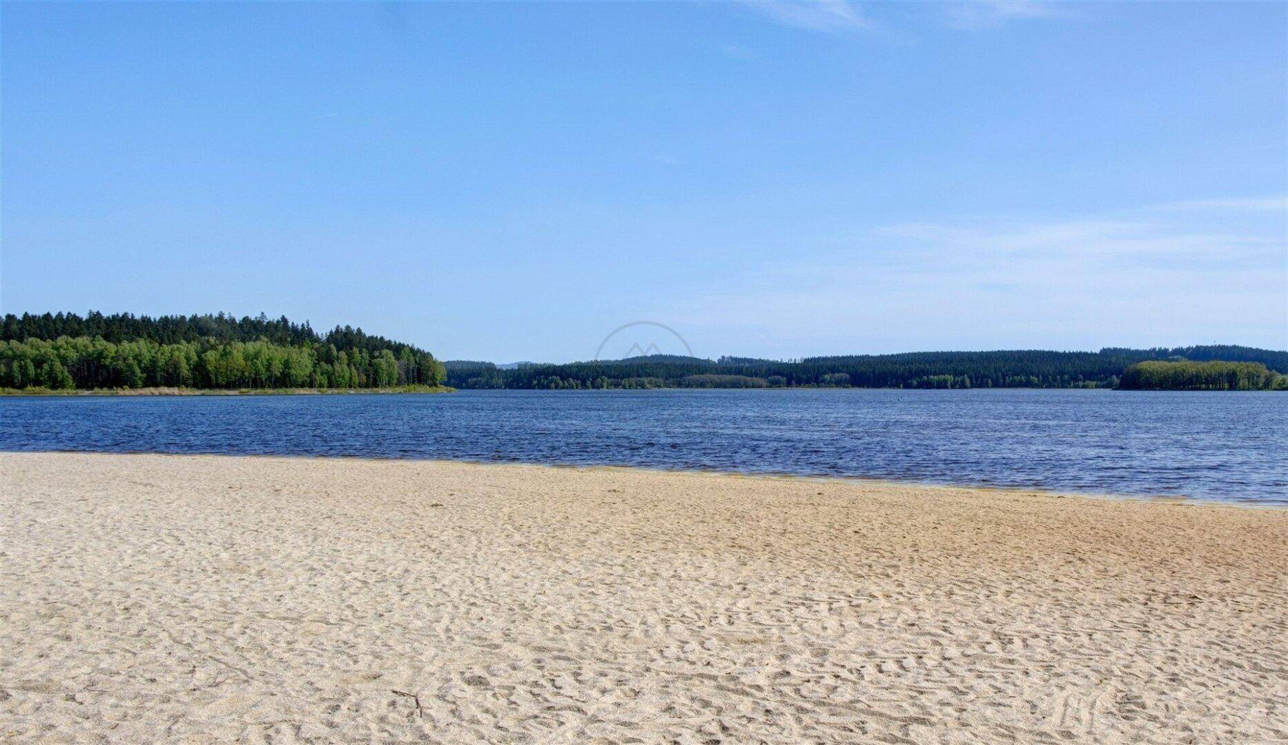Ruhiger Sandstrand am Seeufer mit klarem Wasser und bewaldeten Hügeln im Hintergrund.