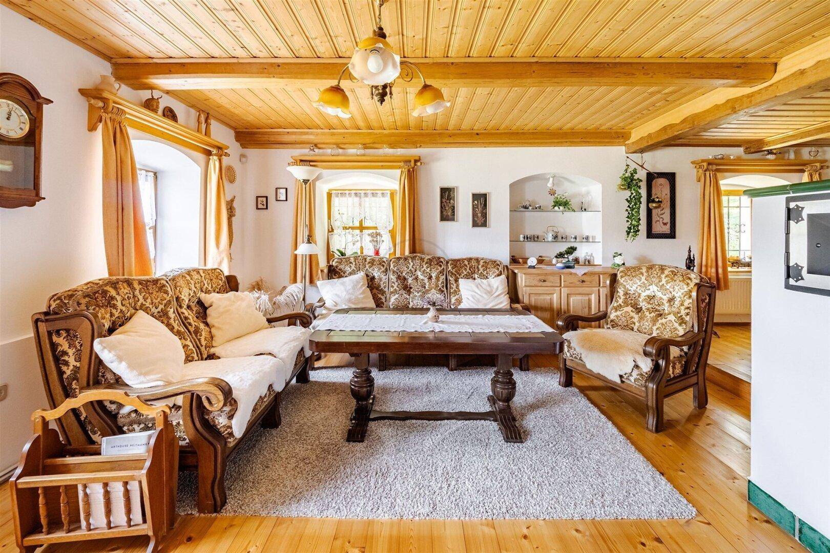 Helles Wohnzimmer mit rustikalen Holzdecken, bequemen Sofas und einem traditionellen Kachelofen im Hintergrund.