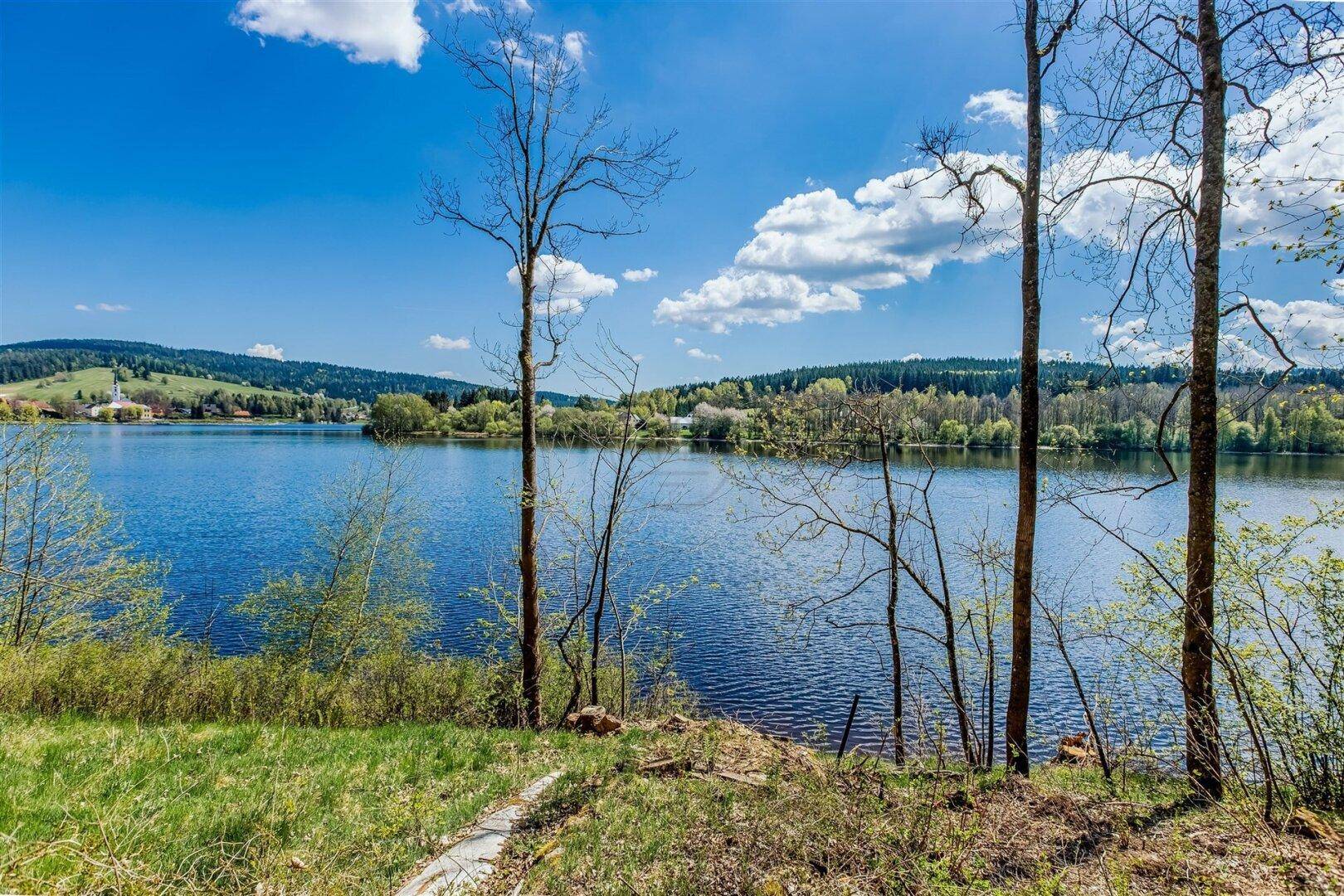 Weitläufiger Blick über einen ruhigen See mit bewaldeten Ufern und blauem Himmel.