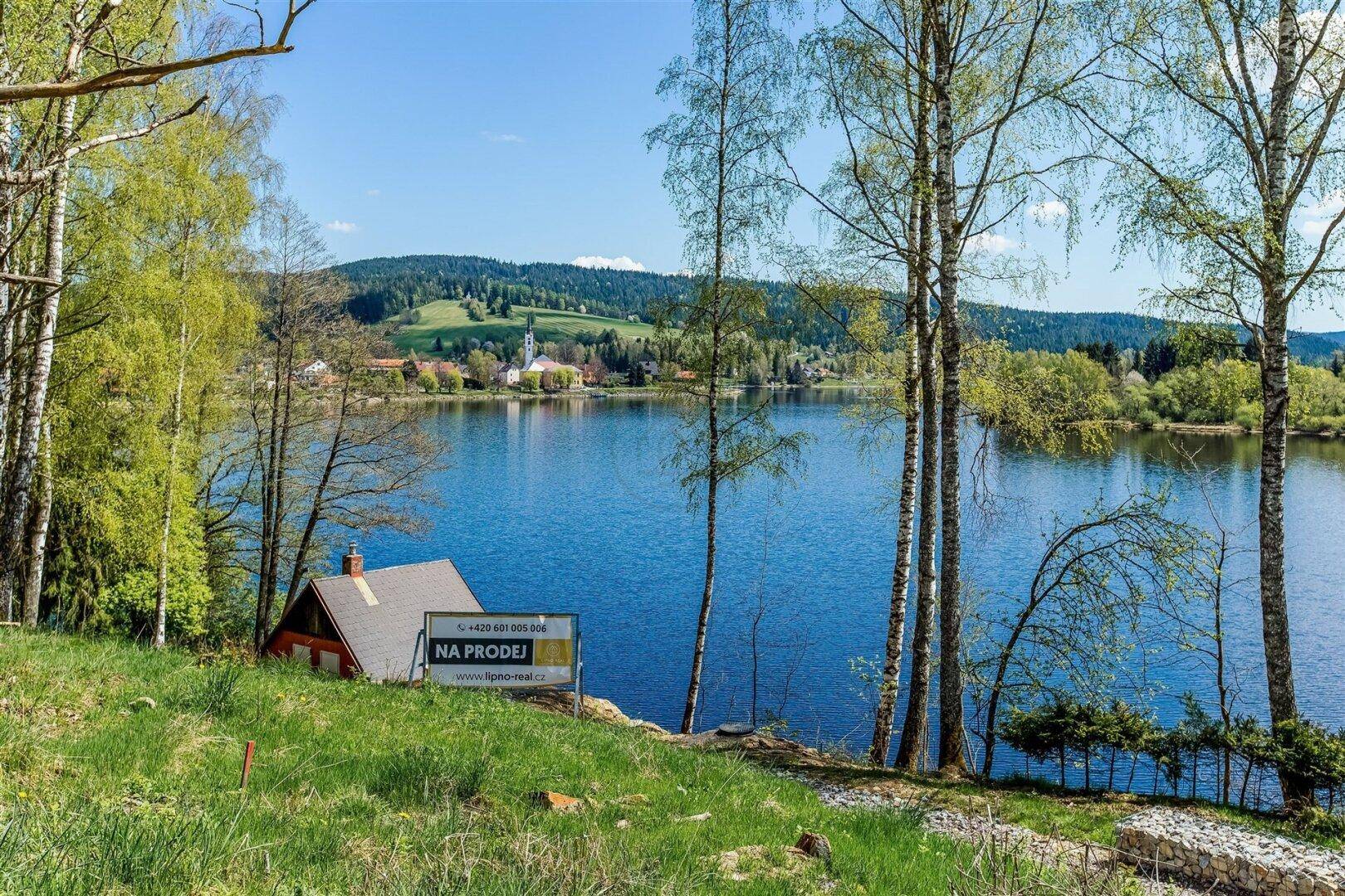 Kleines Haus am Seeufer mit Verkaufsanzeige, umgeben von grüner Landschaft und Bäumen.
