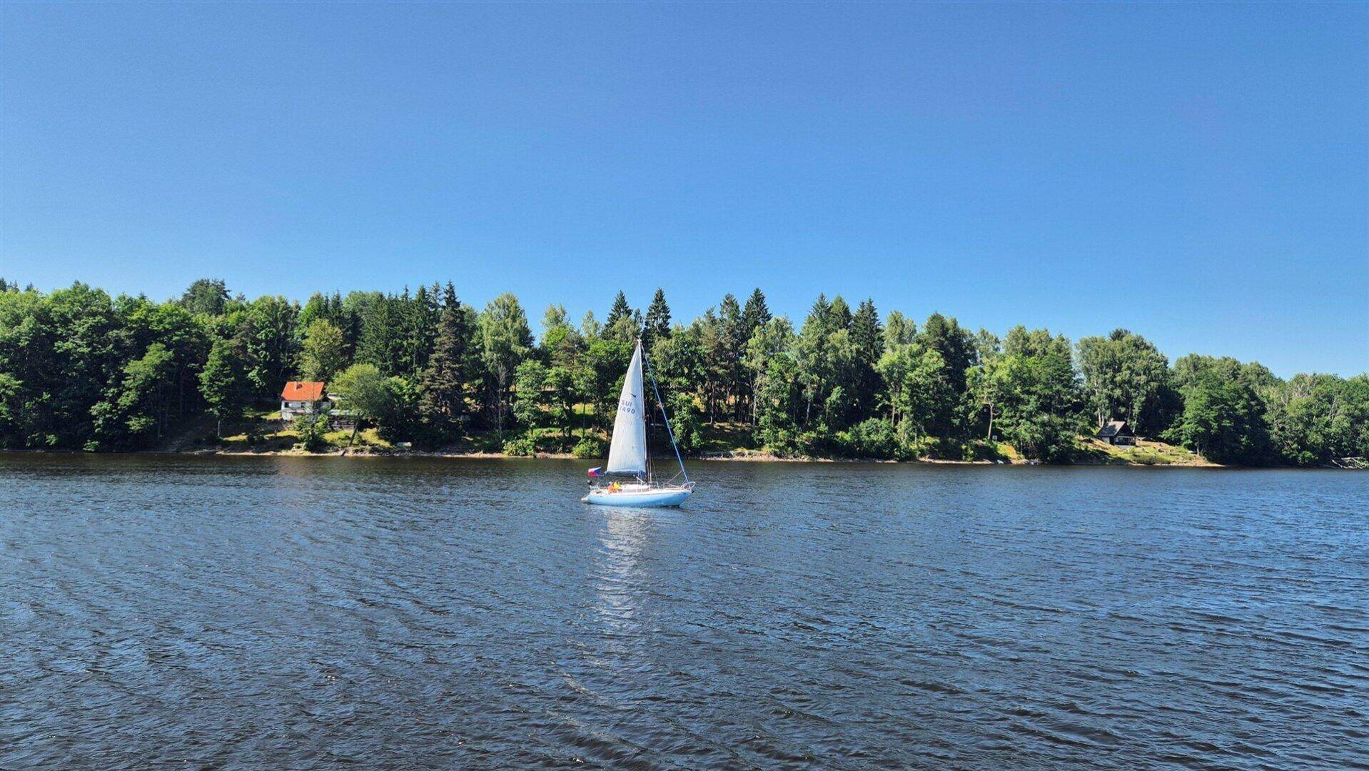 Segelboot auf einem weiten See, umgeben von grünen Wäldern unter klarem Himmel.