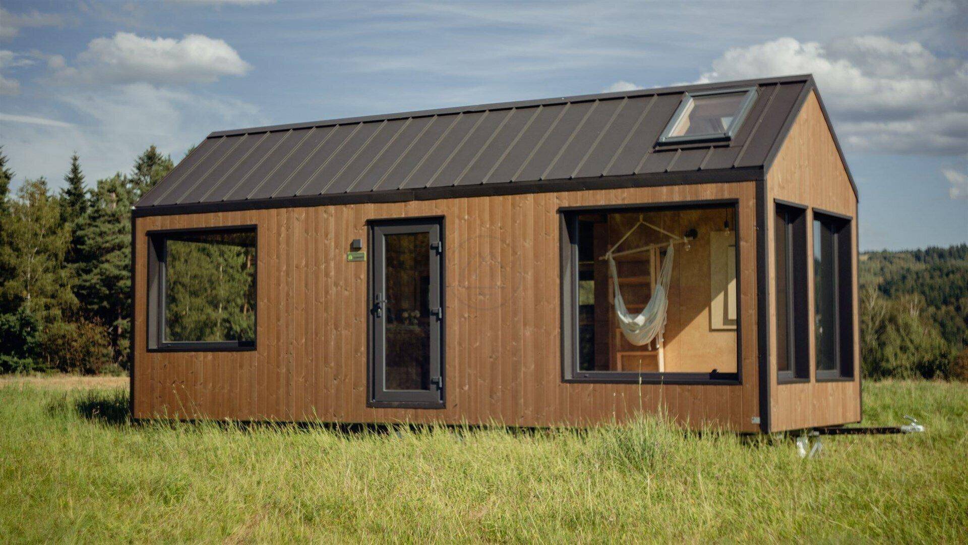 Kompaktes Tiny House mit Holzverkleidung und großen Fenstern, auf Rädern in der Natur.