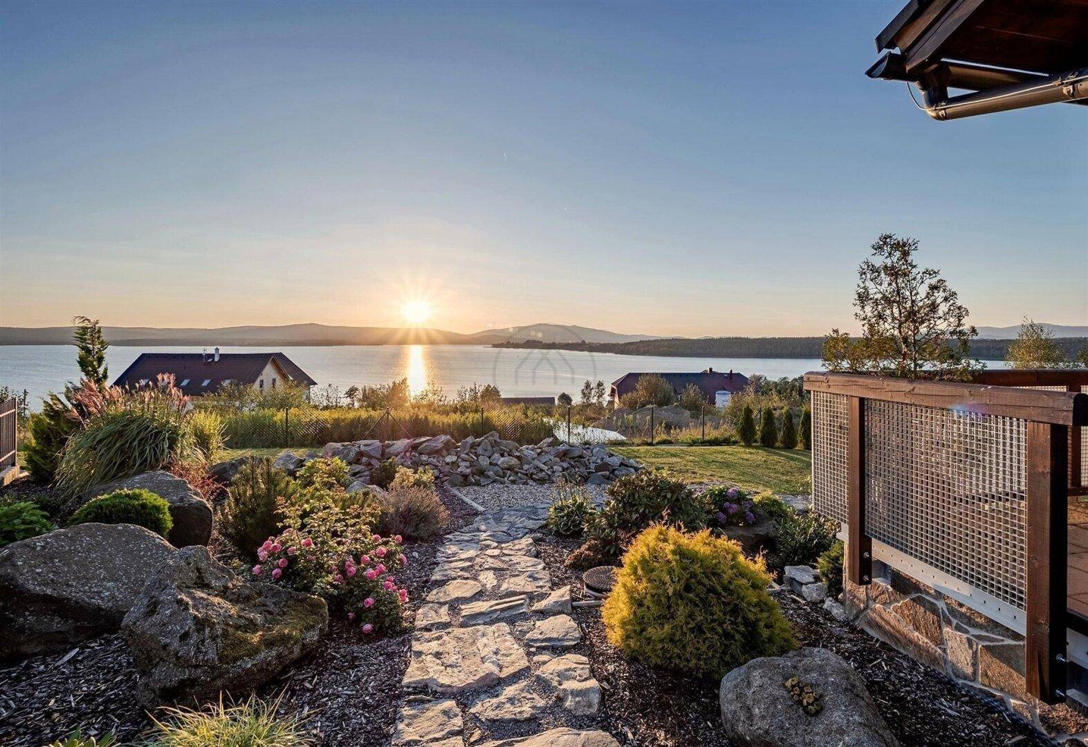 Gartenweg mit Steinplatten, Blumenbeeten und malerischem Blick auf den See bei Sonnenuntergang.
