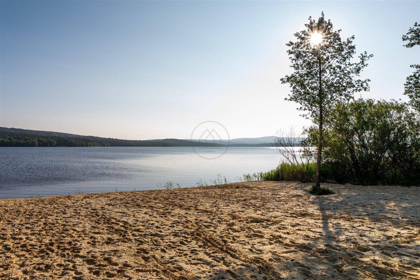 Sandstrand am Seeufer mit klarem Wasser und sonnigem Himmel.
