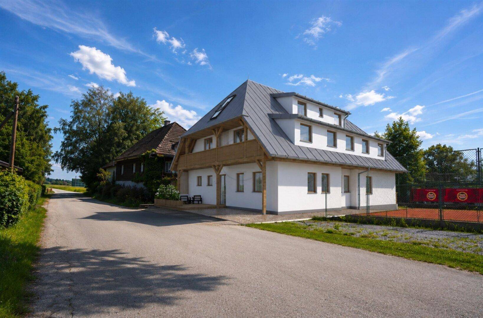 Neubau mit Satteldach und Balkon, umgeben von Natur und einem Tennisplatz.
