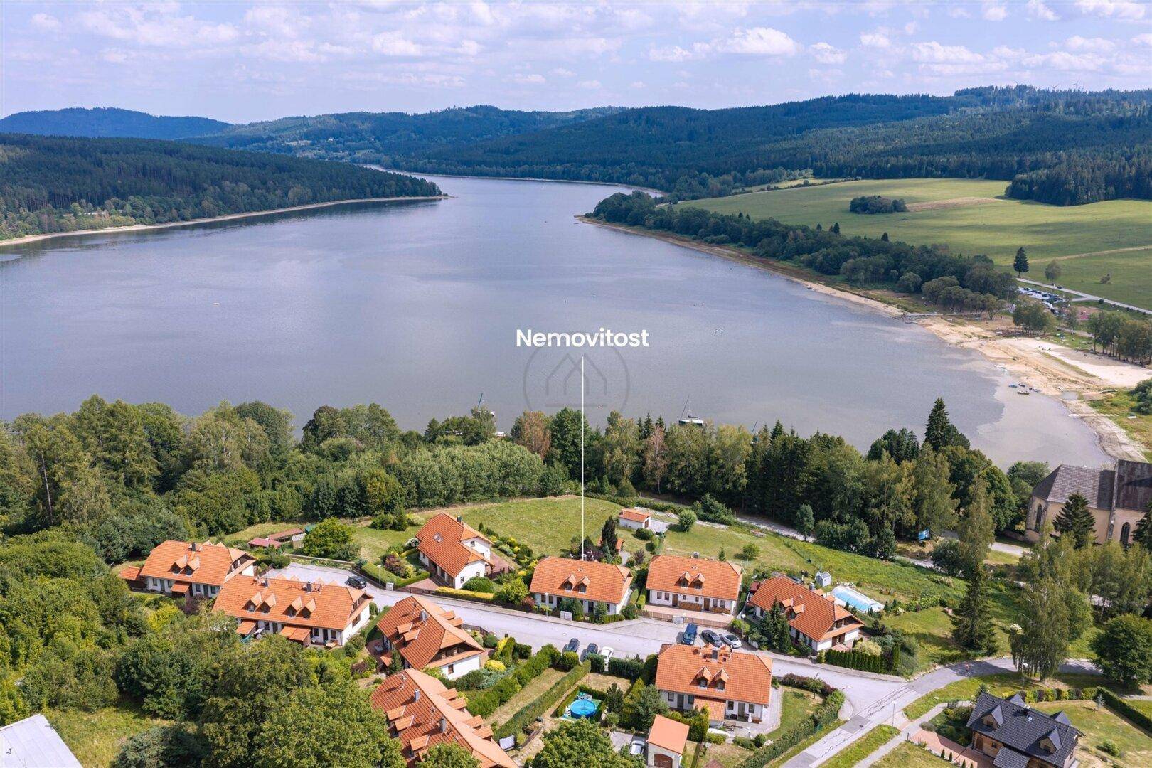 Luftaufnahme einer Wohnsiedlung am Seeufer mit umliegenden Wäldern und Bergen unter blauem Himmel.