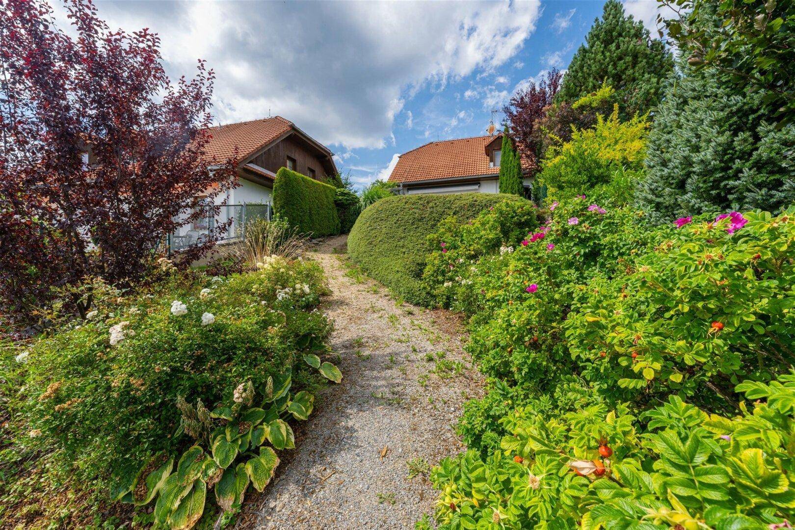 Idyllischer Gartenweg umgeben von üppiger Vegetation und blühenden Sträuchern mit Häusern im Hintergrund.