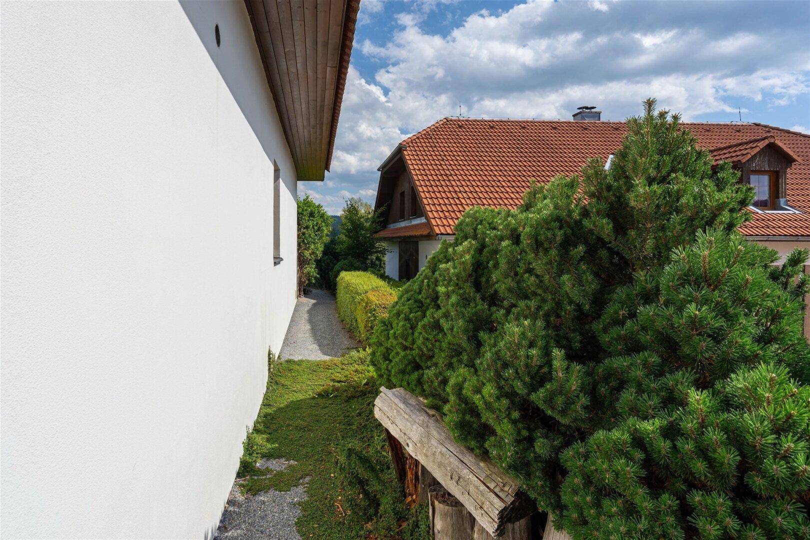 Schmaler Weg entlang eines weißen Hauses mit Blick auf ein weiteres Gebäude und grüne Hecken.