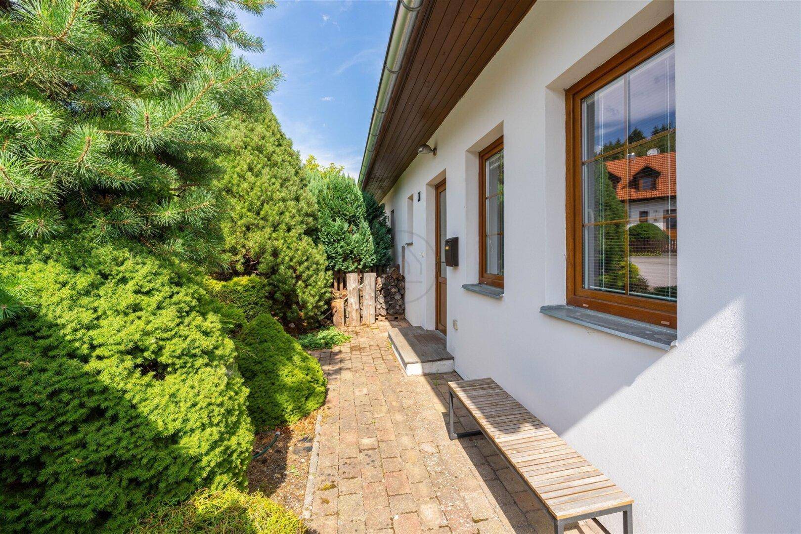 Seitlicher Zugang zum Haus mit gepflastertem Weg, üppiger Bepflanzung und einer Holzbank.