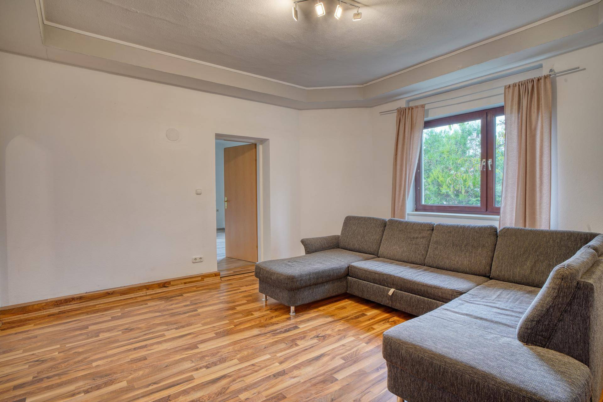 Helles Wohnzimmer mit Parkettboden, großer Eckcouch und Fenster mit Vorhängen.