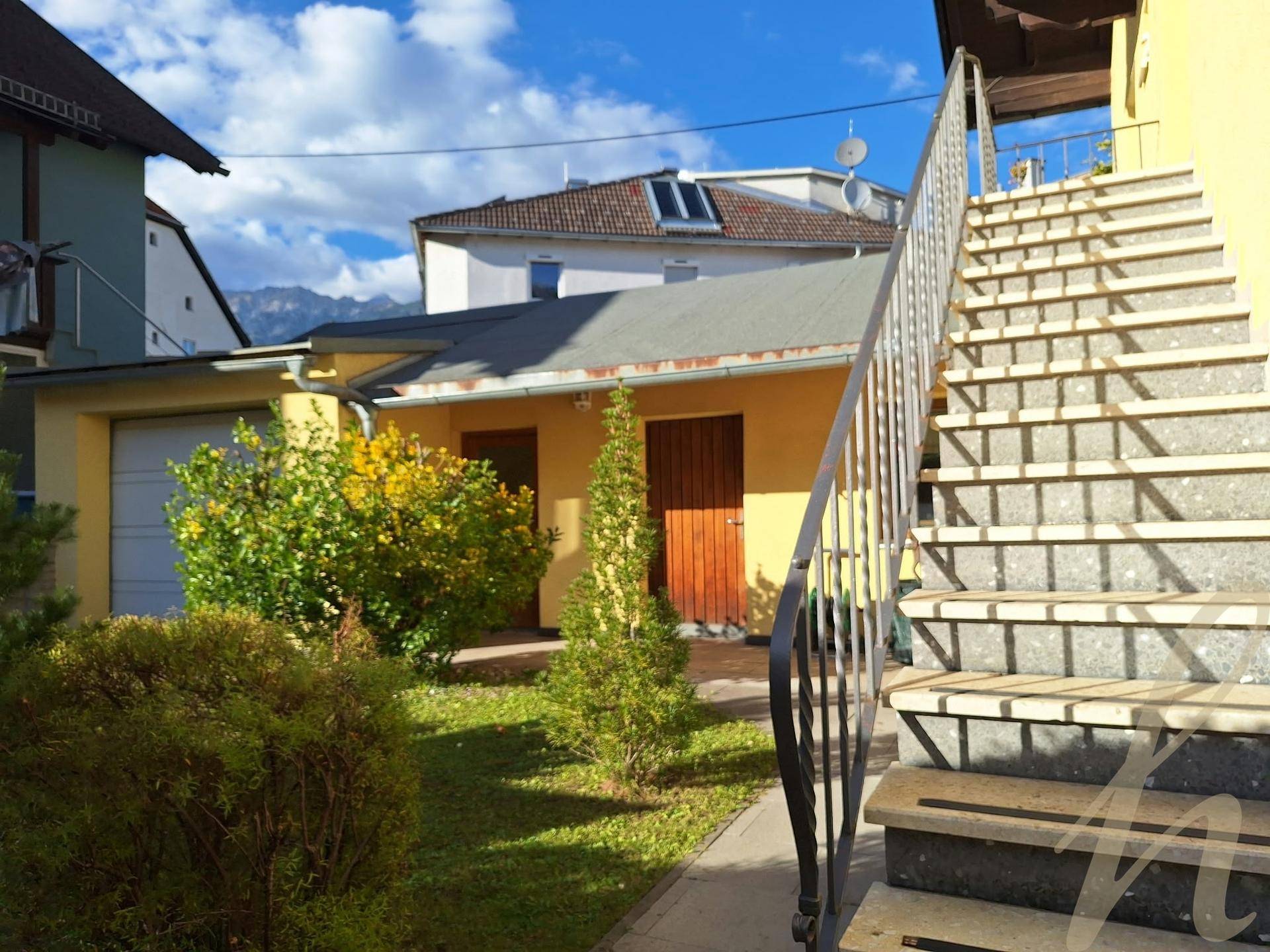 Außenansicht des gelben Hauses mit Gartenbereich, Garage und Blick auf die Berge.
