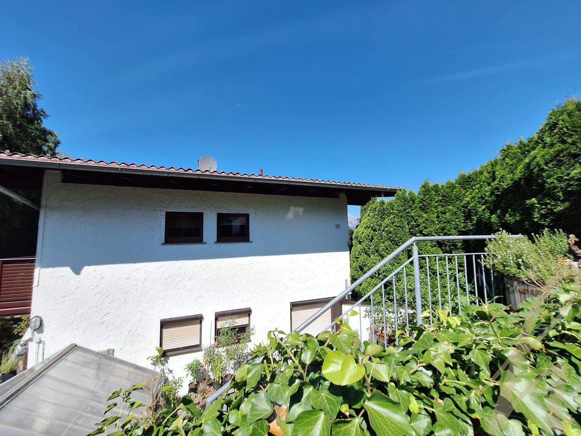 Seitenansicht des Hauses mit Außentreppe und üppiger grüner Bepflanzung unter blauem Himmel.