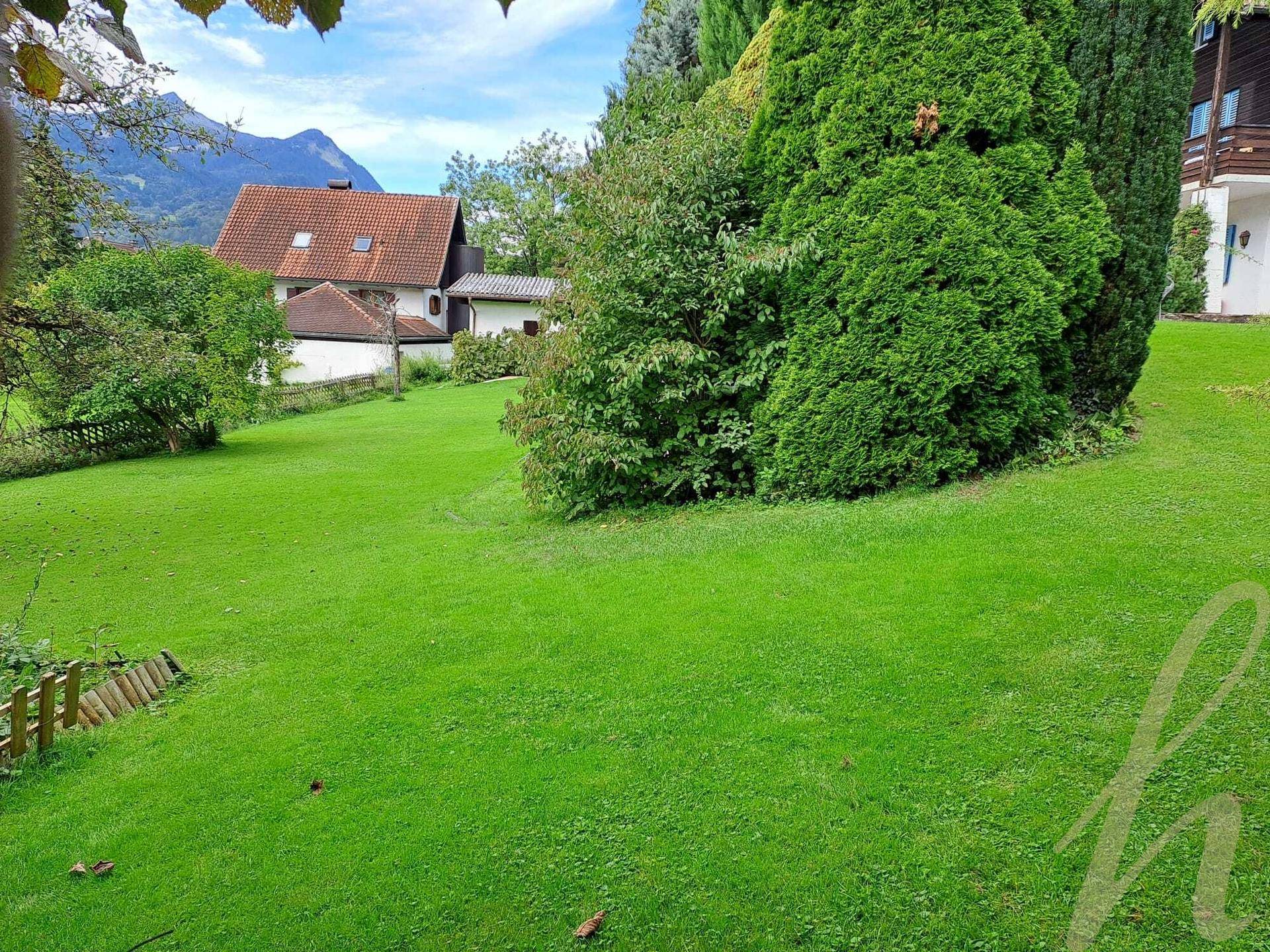 Weitläufiger Rasenbereich im Garten mit Blick auf traditionelle Häuser und die majestätischen Berge im Hintergrund.