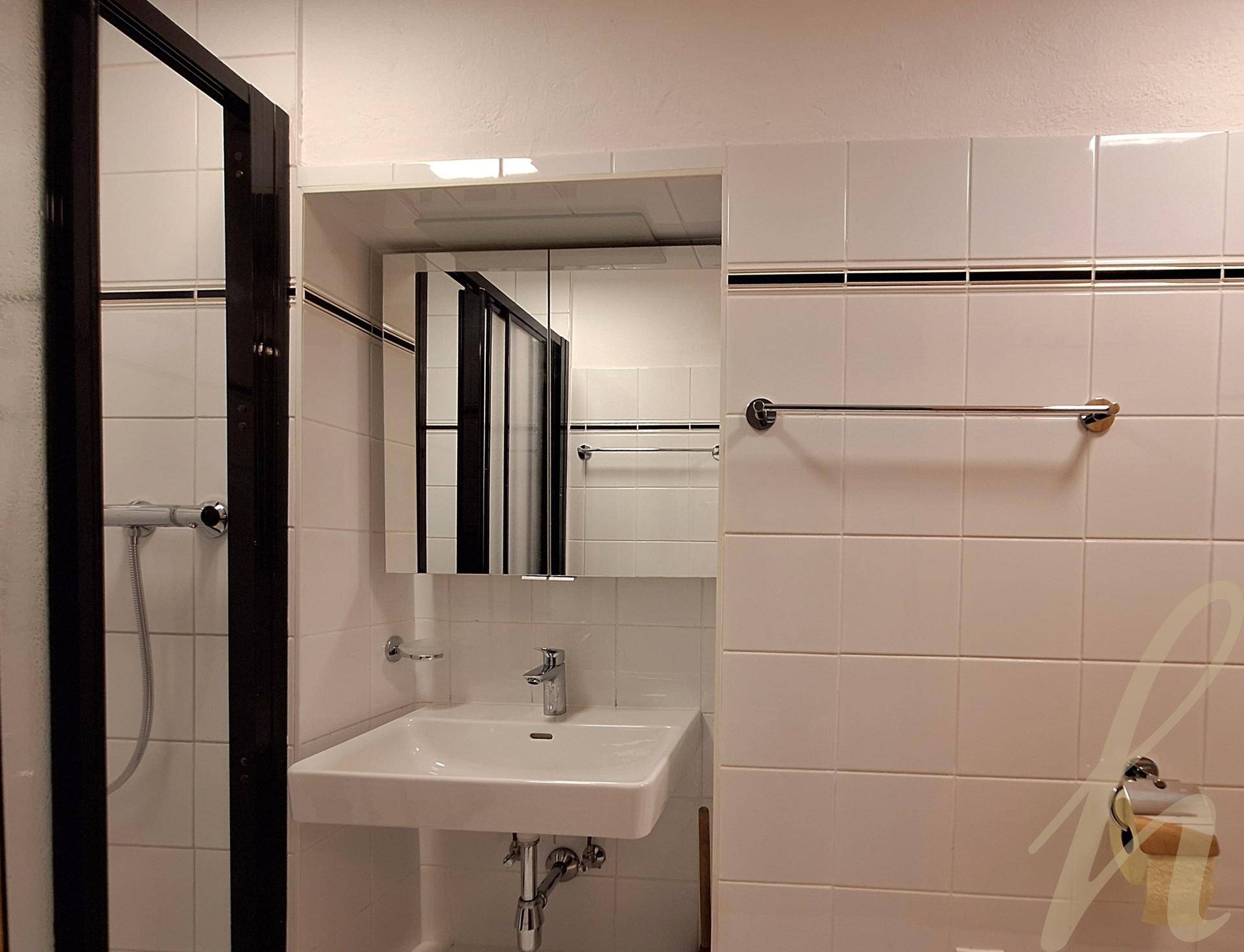 Funktionales Badezimmer mit weißen Fliesen, einer Dusche und einem Waschbecken mit Spiegelschrank.