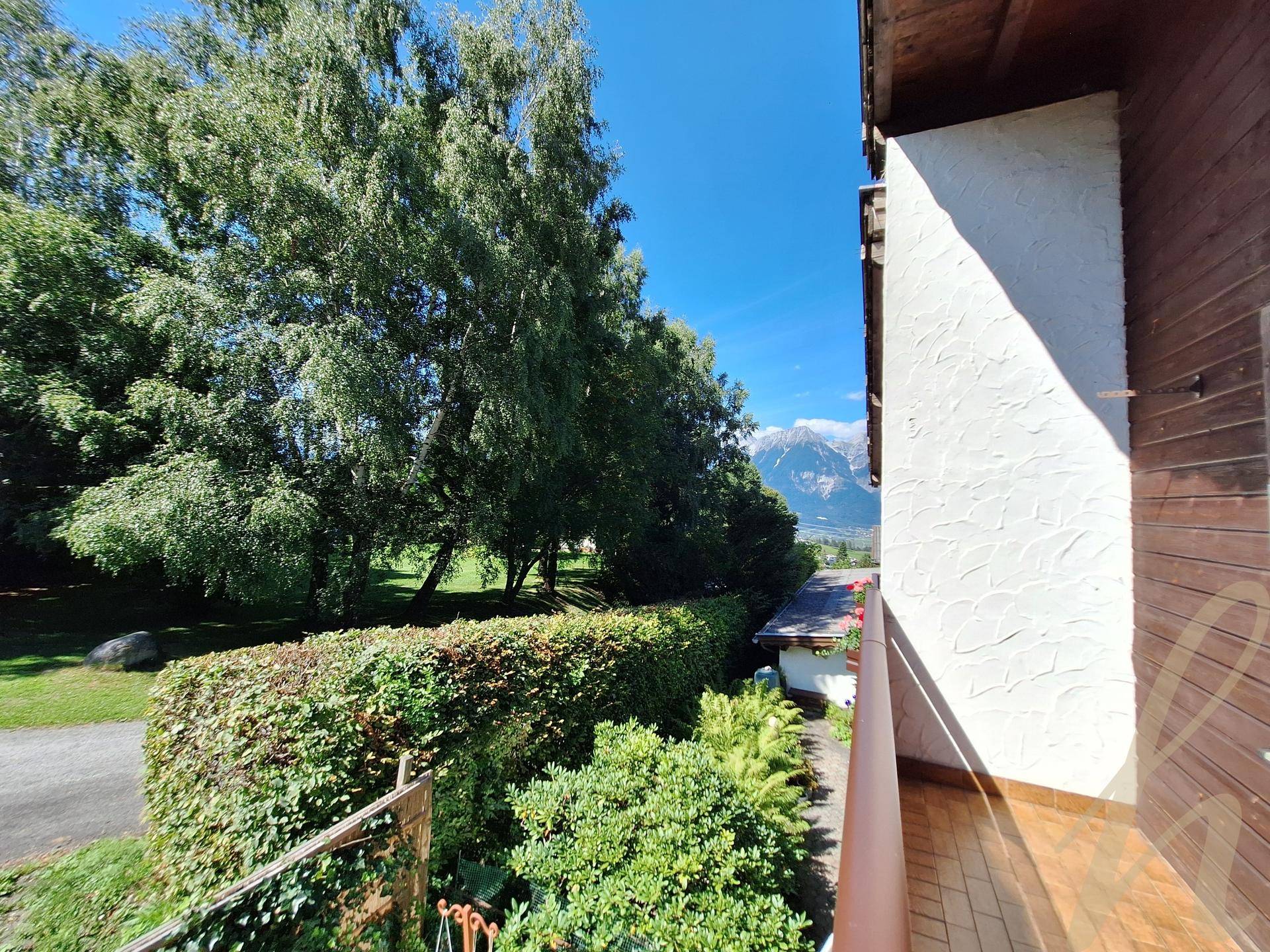 Weitblick vom Balkon über eine grüne Hecke und Bäume bis zu den majestätischen Bergen.