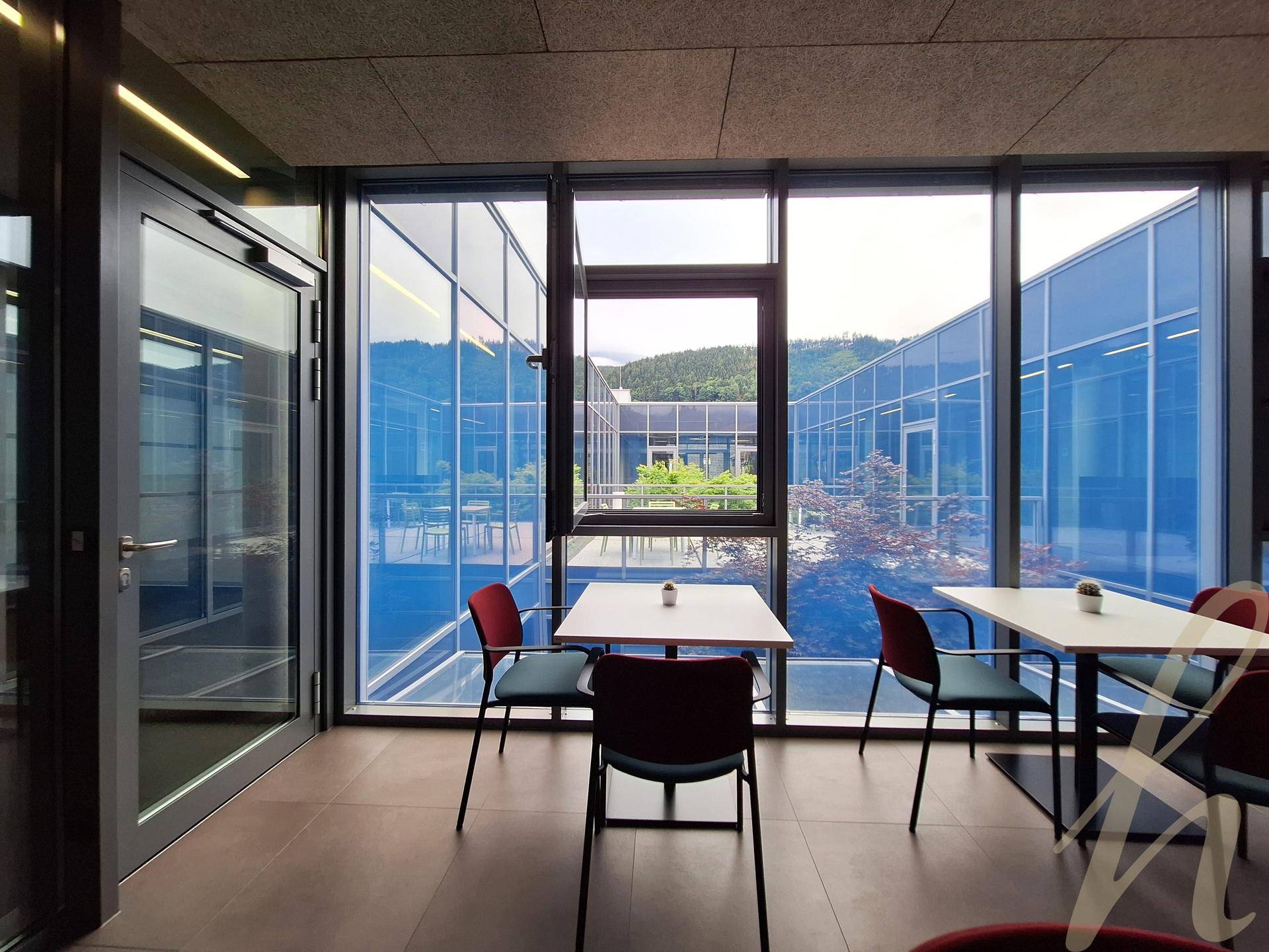 Helle Cafeteria mit kleinen Tischen und Stühlen, umgeben von Glaswänden mit Blick auf den Innenhof und grüne Hügel.