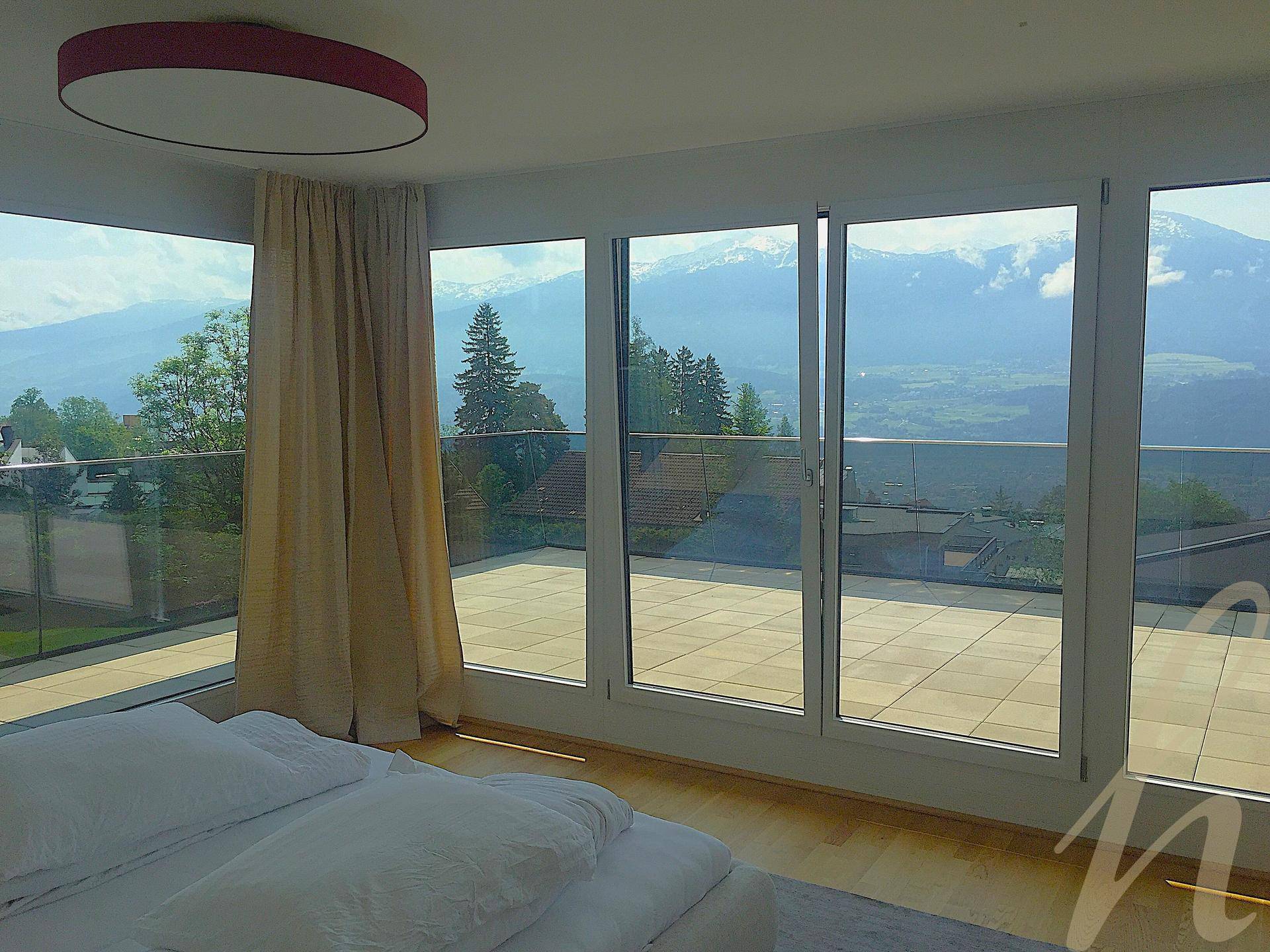 Schlafzimmer mit großem Bett, Holzboden und raumhohen Fenstern, die Zugang zur Terrasse und Bergblick bieten.