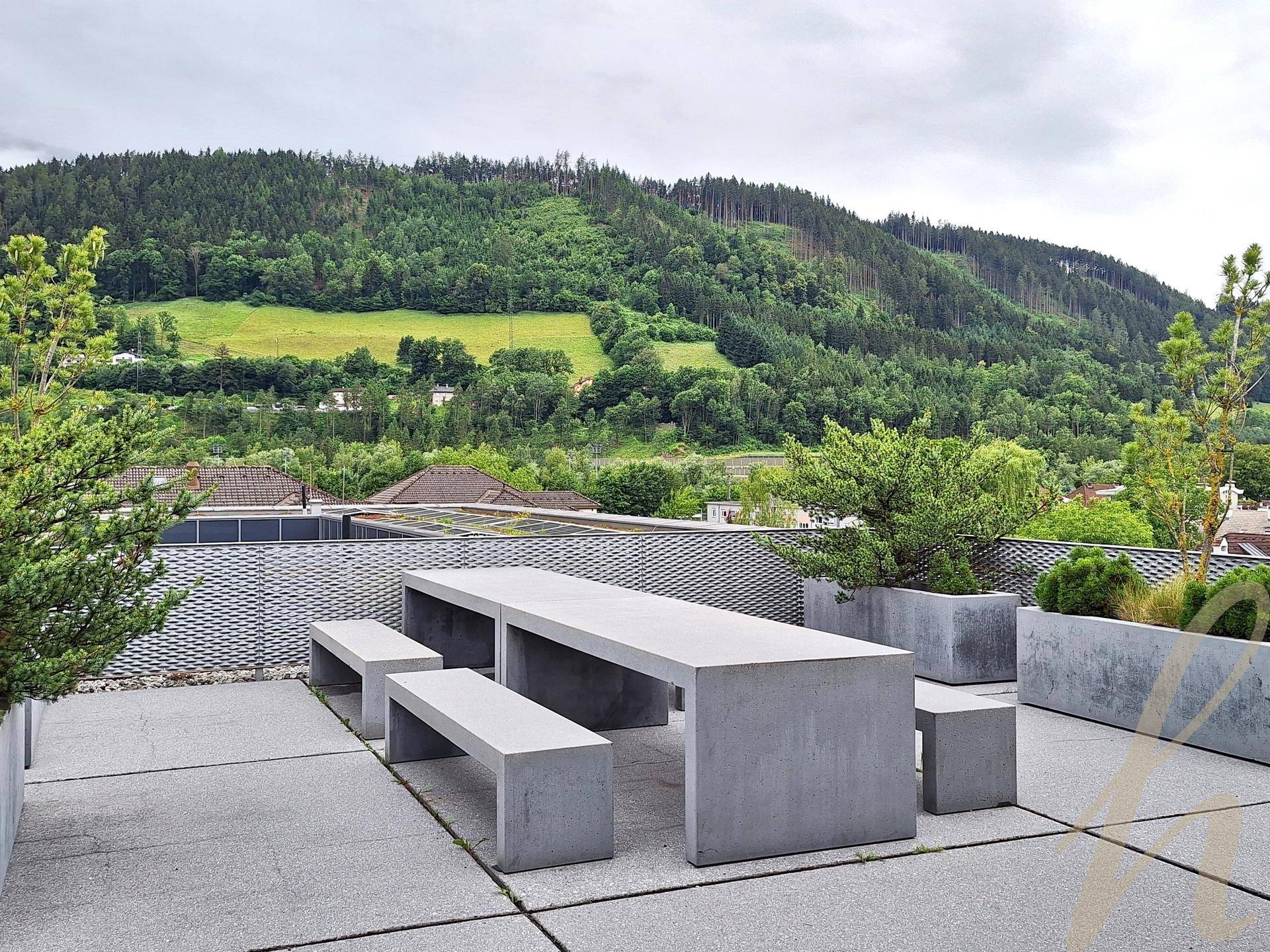 Weitläufige Dachterrasse mit modernen Betonmöbeln und Pflanzkübeln, umgeben von grünen Hügeln und Bergen.
