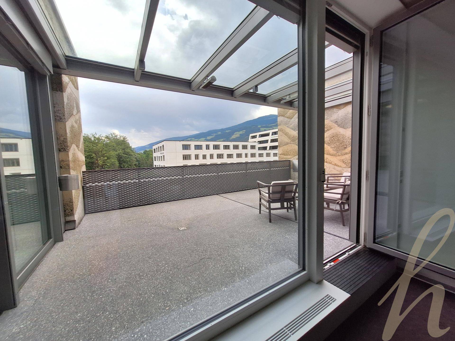 Geräumige Terrasse mit Glasdach, Steinwänden und Sitzgelegenheiten, bietet Blick auf Gebäude und Berge.
