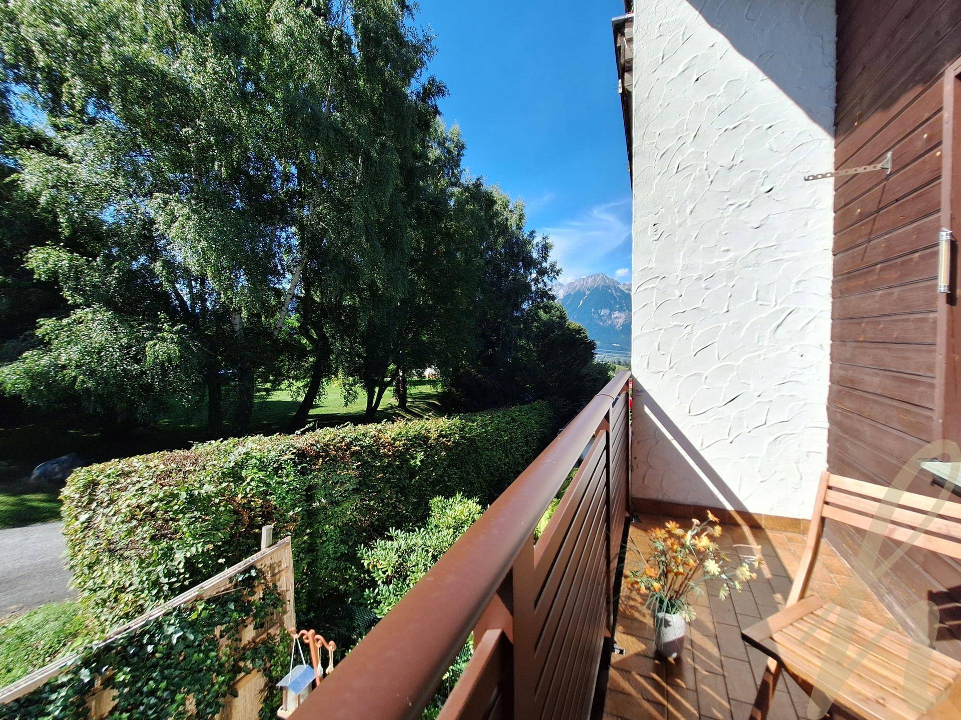 Blick vom Balkon auf eine grüne Hecke, hohe Bäume und die umliegende Berglandschaft.