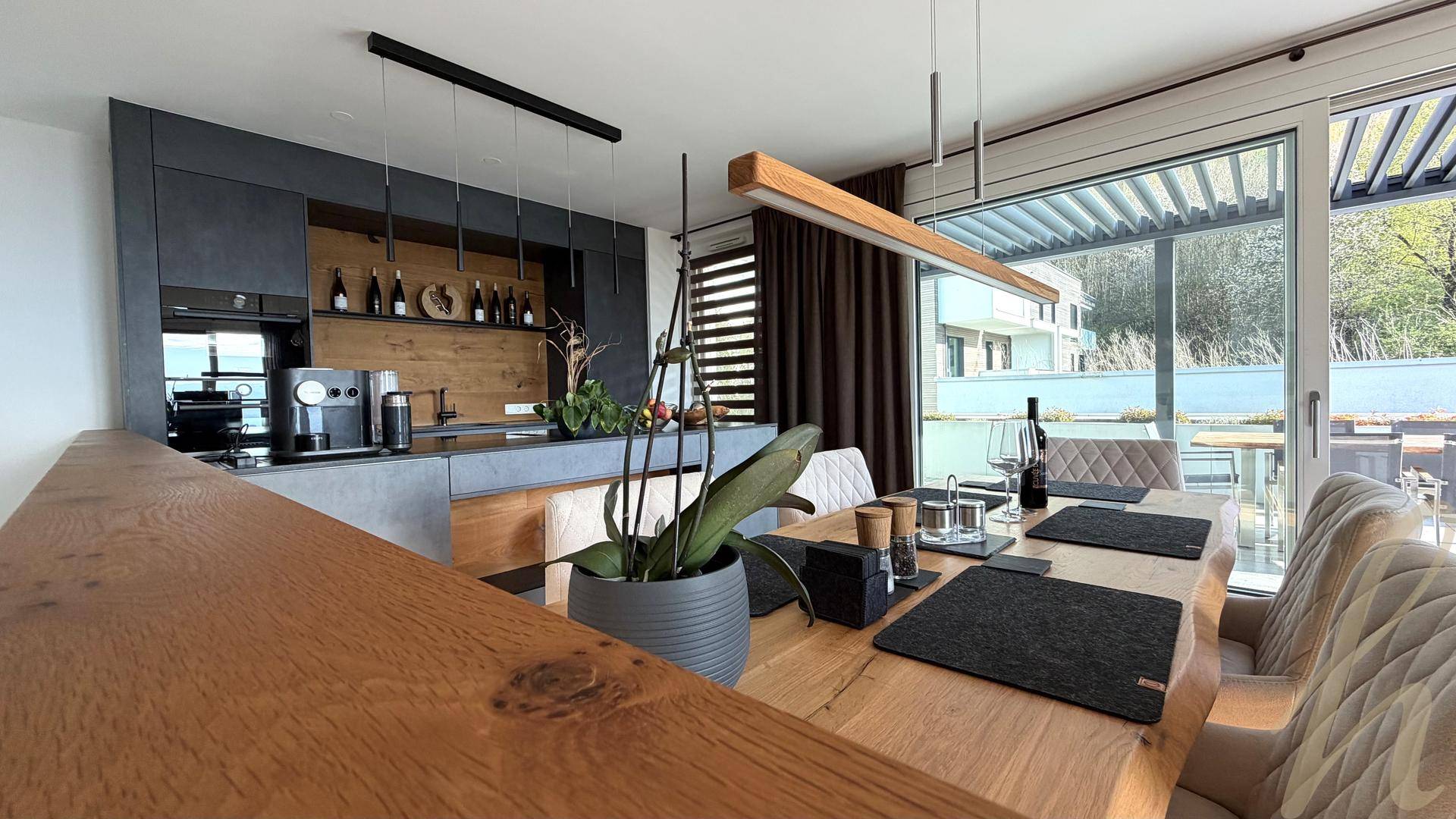Essbereich mit Holztisch und moderner Küche, direkter Zugang zur überdachten Außenterrasse.
