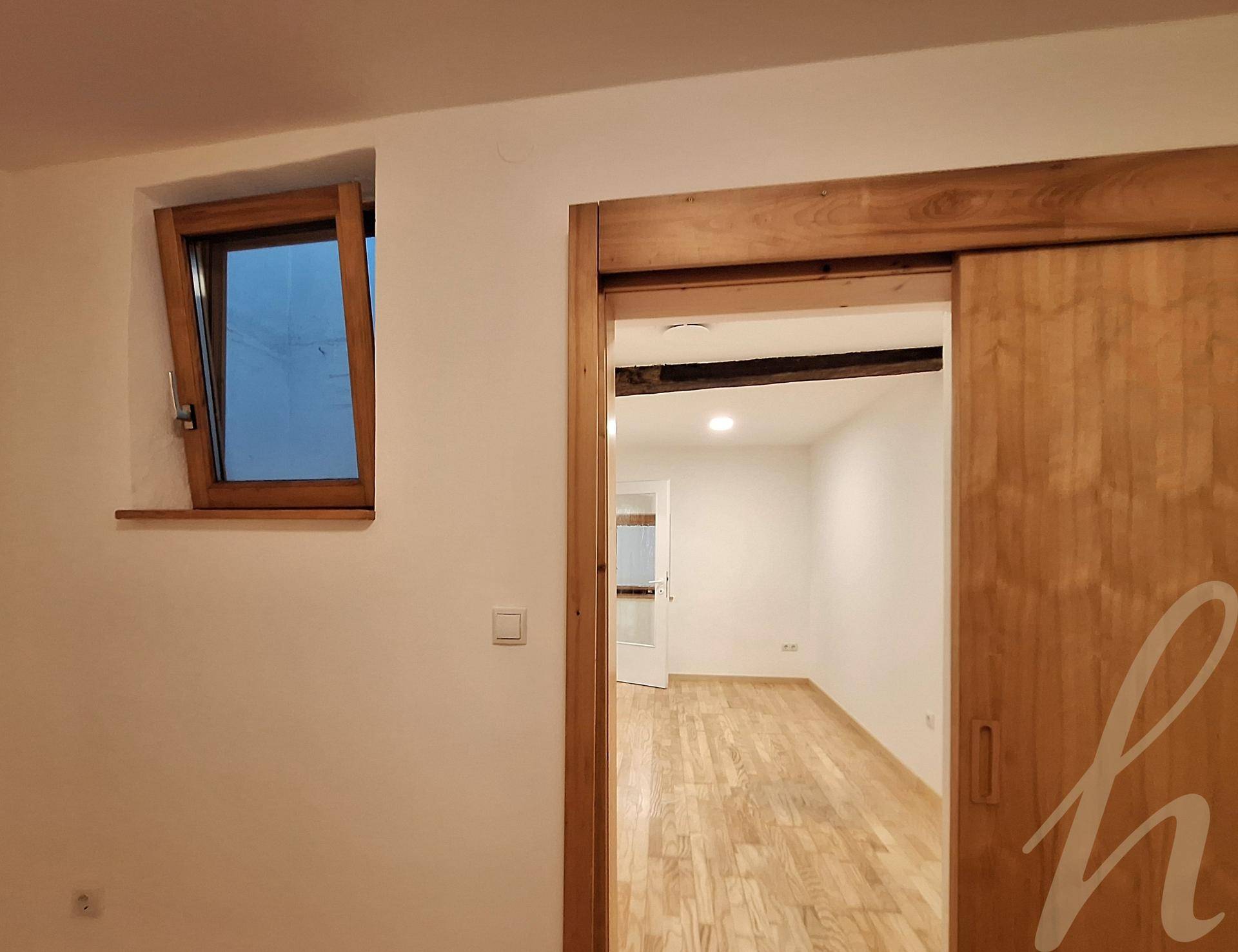 Innenansicht mit einem kleinen Holzfenster, einer Schiebetür und einem Blick auf einen Raum mit Holzboden.