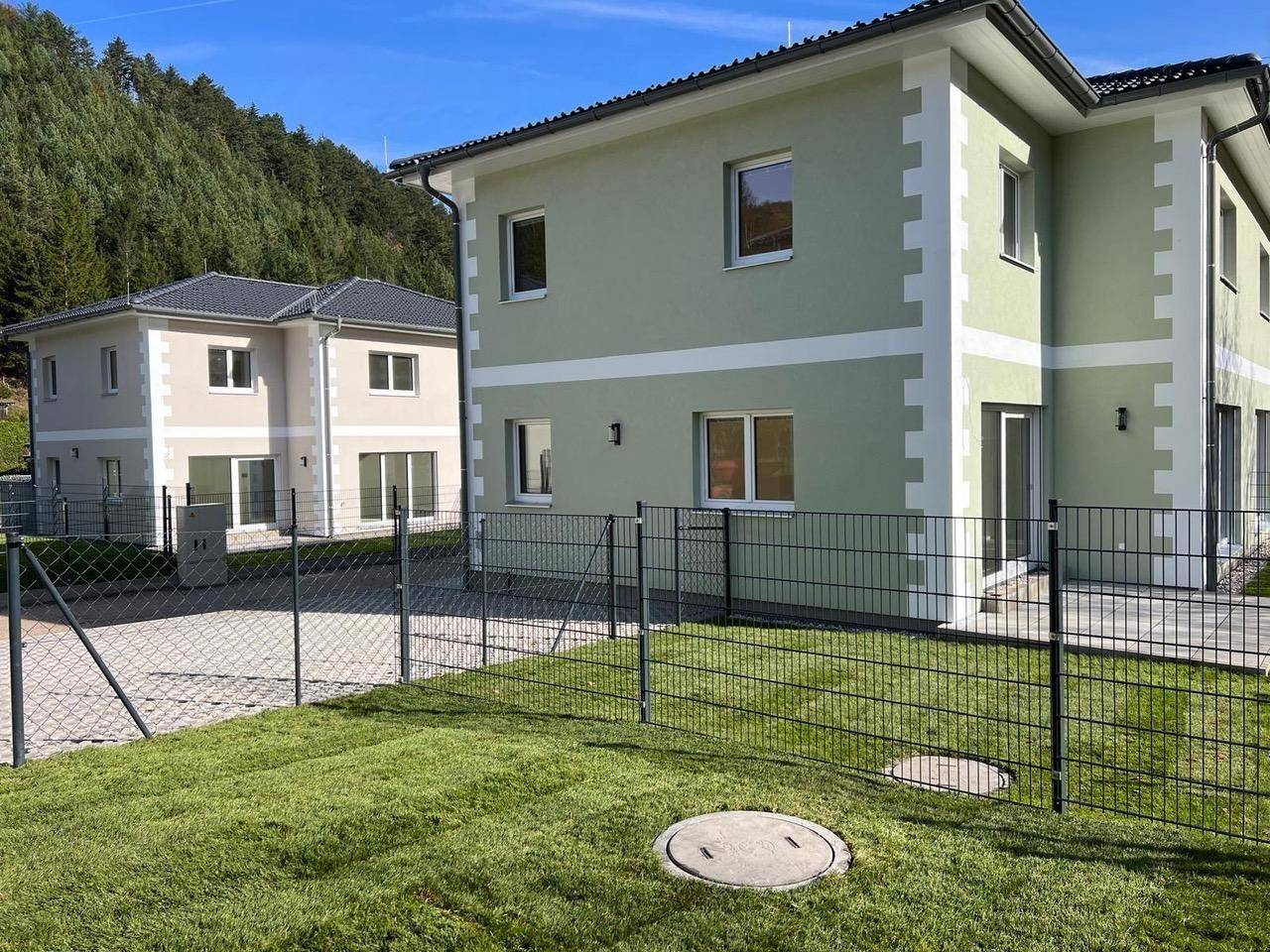 Zwei moderne Neubauten mit hellgrünen Fassaden und umzäunten Gärten in sonniger Lage.