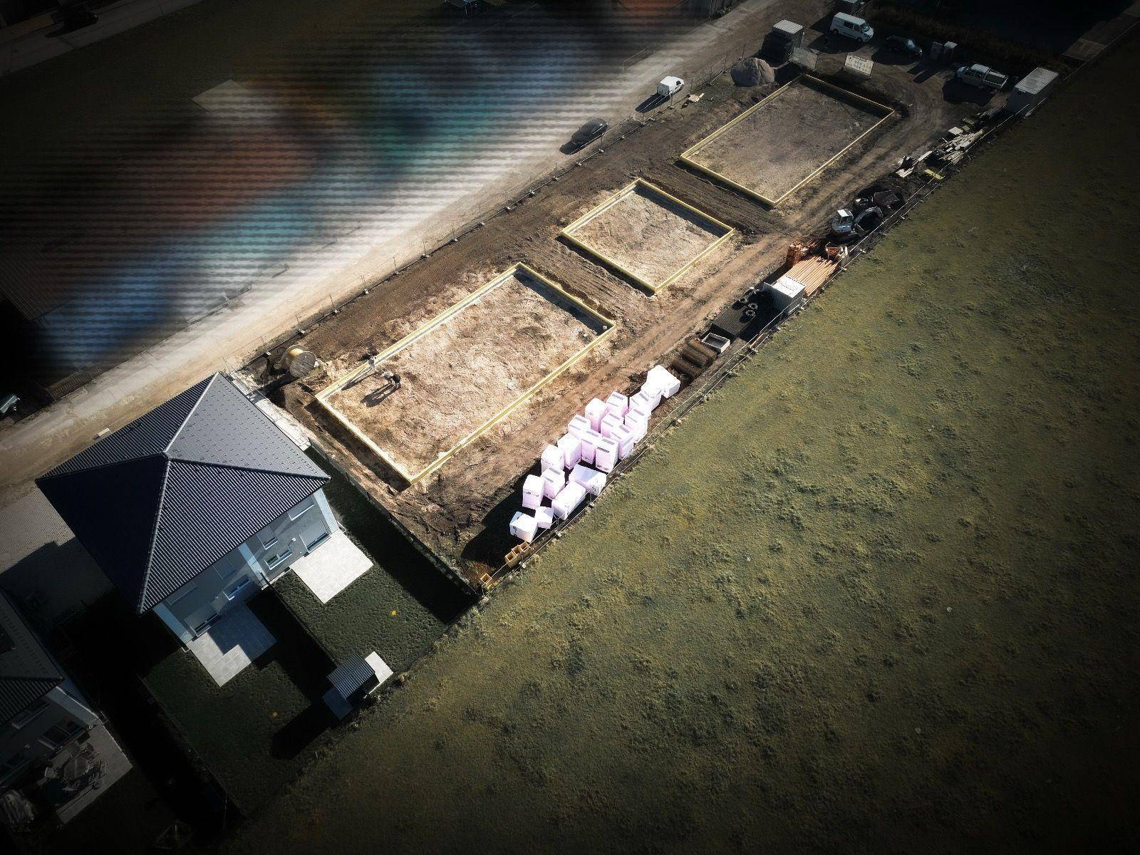 Schräge Luftaufnahme einer Baustelle mit Fundamenten und einem bereits fertiggestellten Haus.