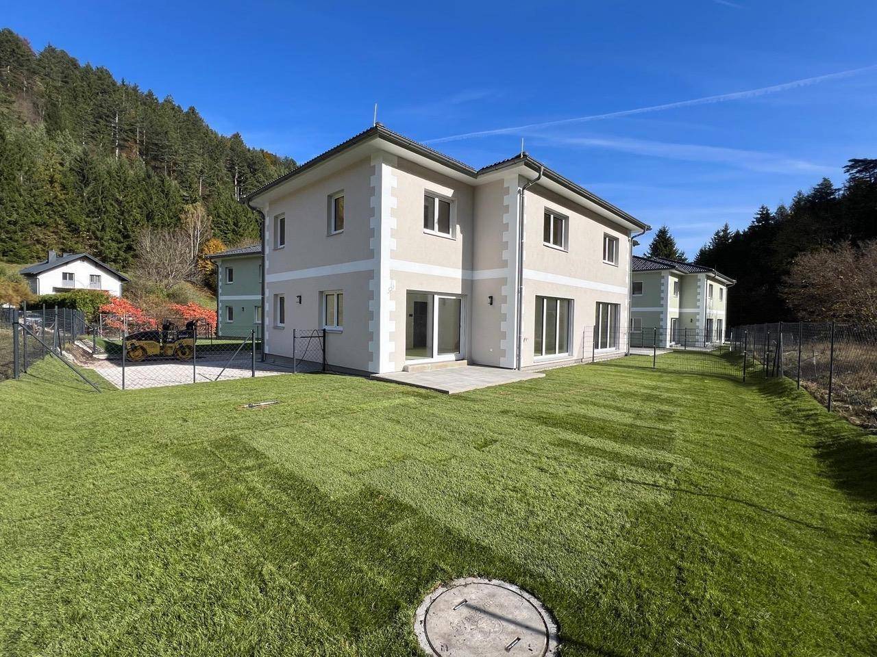 Neubau mit großem, frisch angelegtem Rasen und Blick auf bewaldete Hügel unter blauem Himmel.