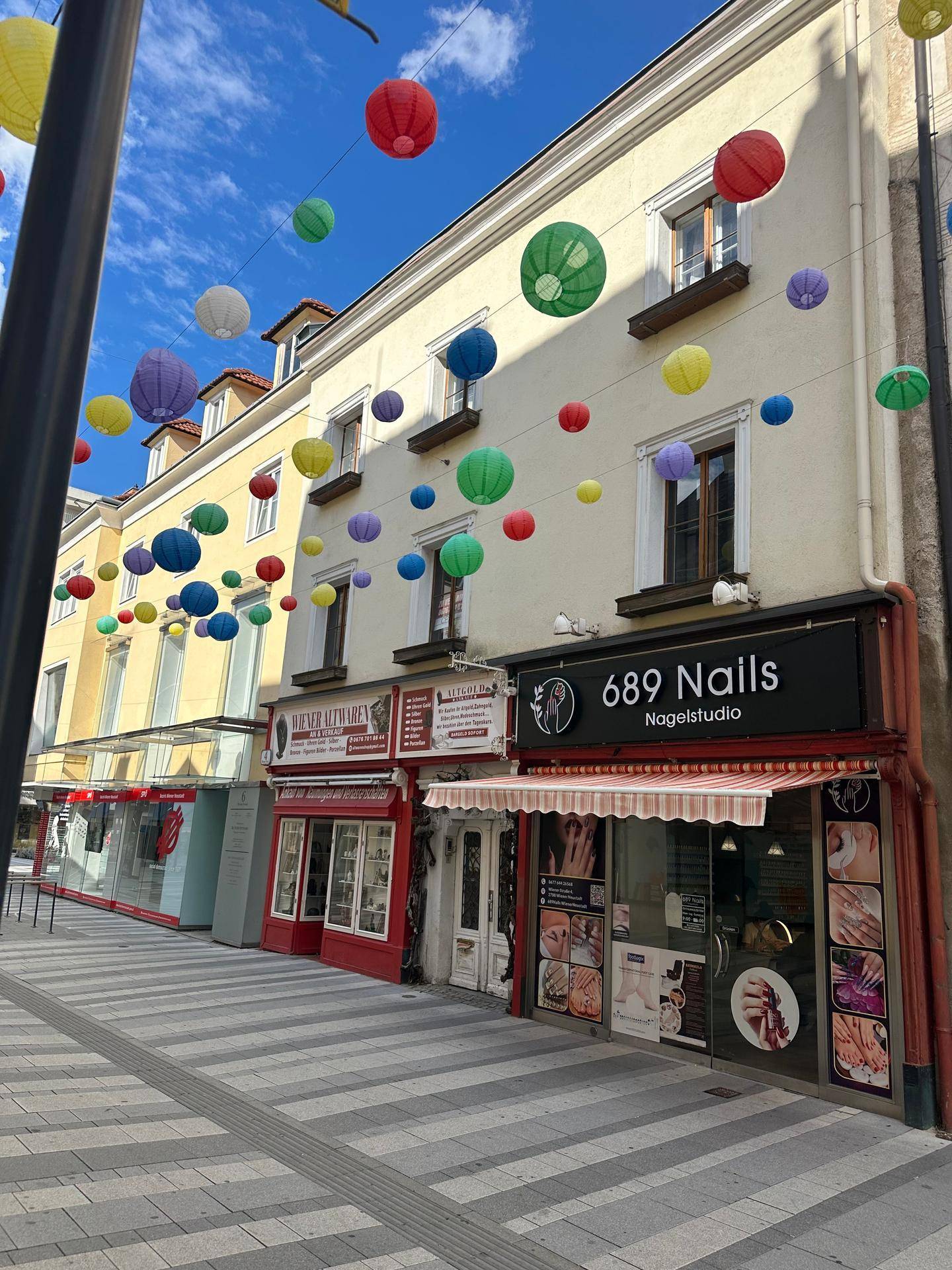 Nahaufnahme einer Geschäftsfassade in einer geschmückten Fußgängerzone mit bunten Laternen unter blauem Himmel.