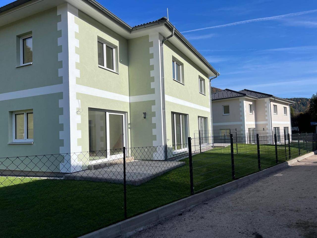 Modernes Haus mit hellgrüner Fassade, großen Terrassentüren und umzäuntem Gartenbereich.