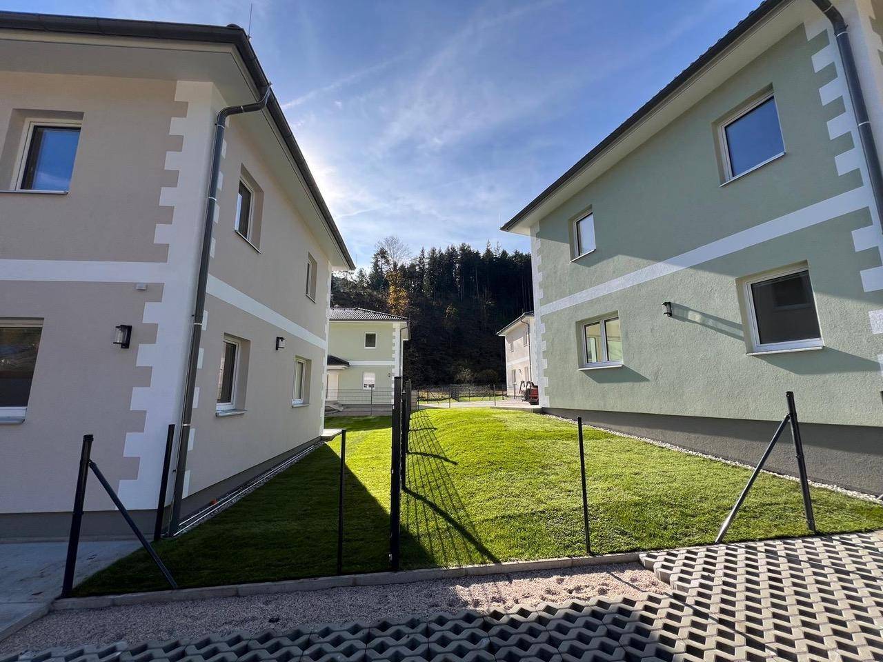 Blick zwischen zwei modernen Häusern mit grünen Rasenflächen und bewaldetem Hintergrund.