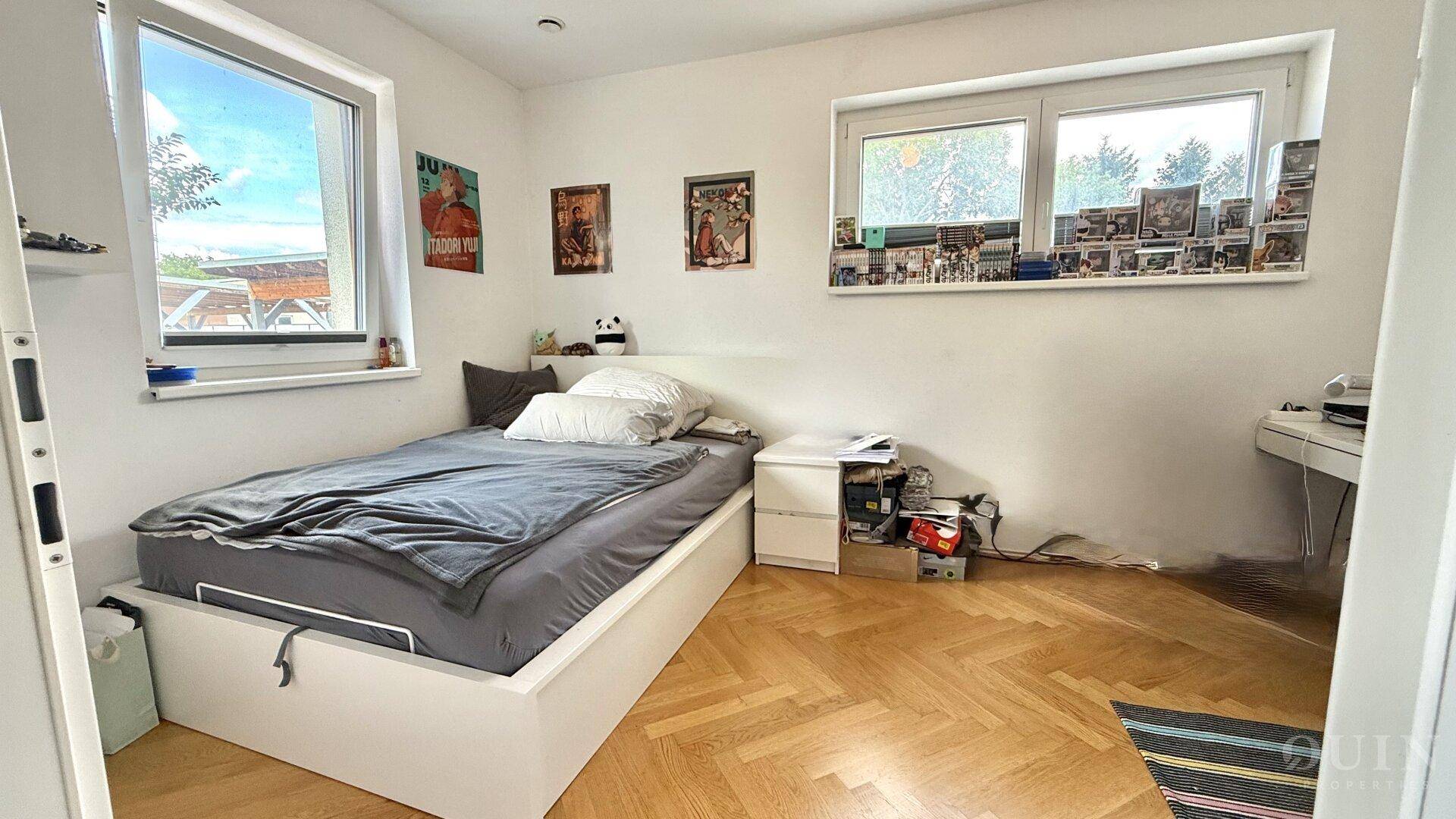 Helles Schlafzimmer mit Bett, Nachttisch und Fenstern, dekoriert mit Postern.