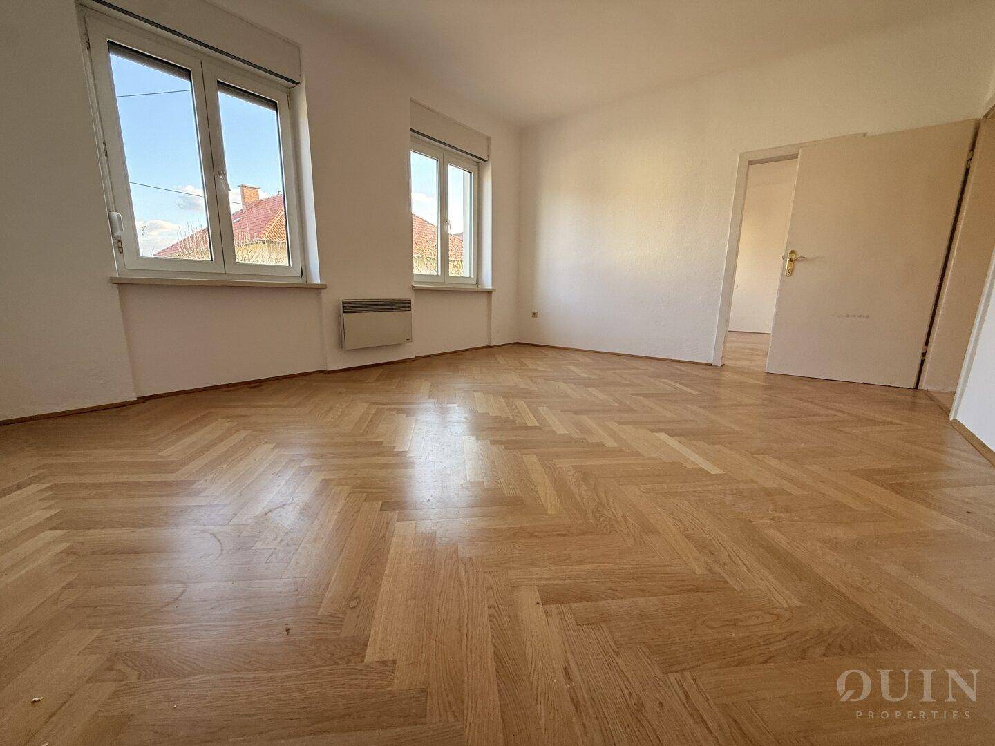 Schlafzimmer mit Fischgrätparkett und zwei Fenstern, die viel Tageslicht hereinlassen.