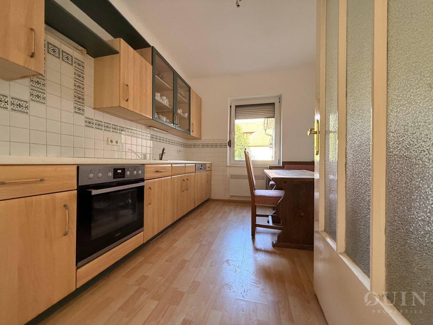 Funktionale Küche mit Holzschränken, Einbauherd und Essbereich am Fenster.