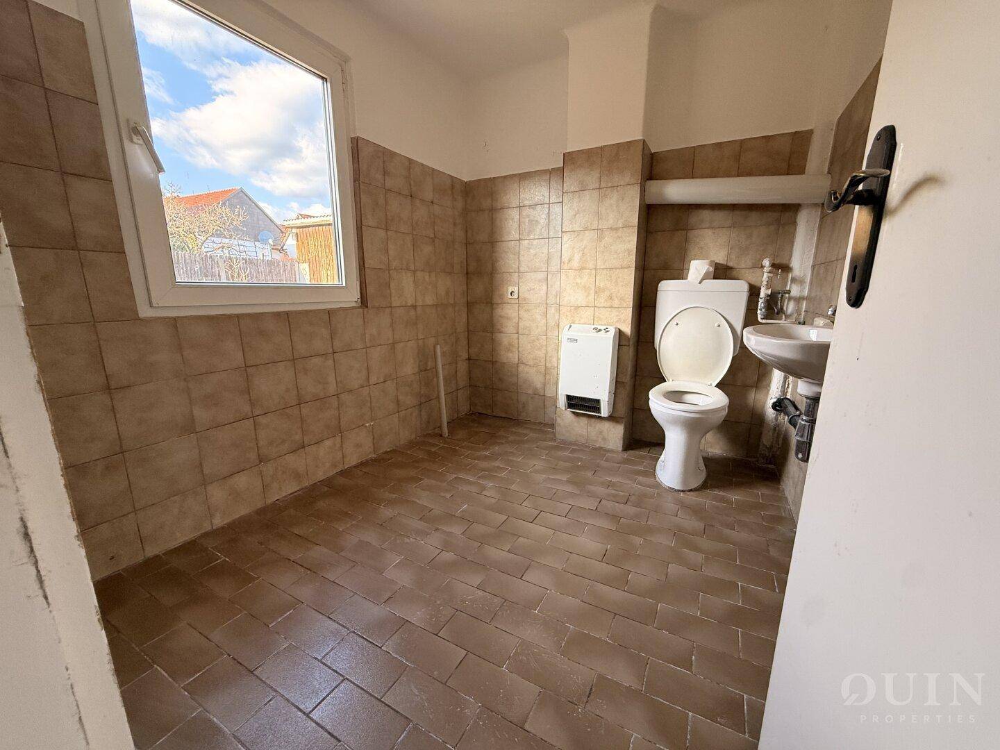 Einfaches WC mit Fenster, Toilette und Waschbecken, gefliest in Brauntönen.