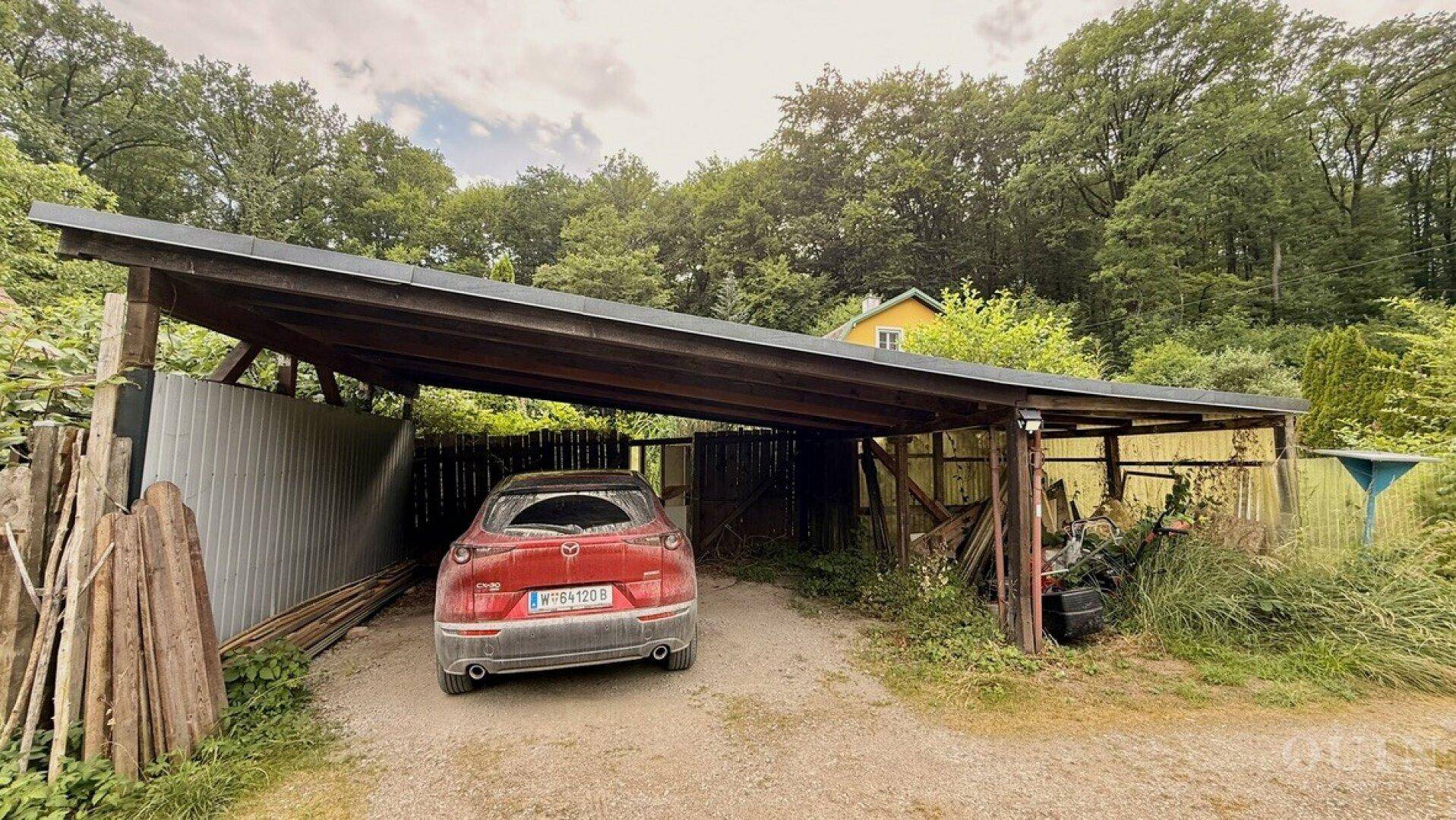 Holz-Carport mit einem geparkten Auto, umgeben von üppiger Vegetation und einem Zaun im Grünen.