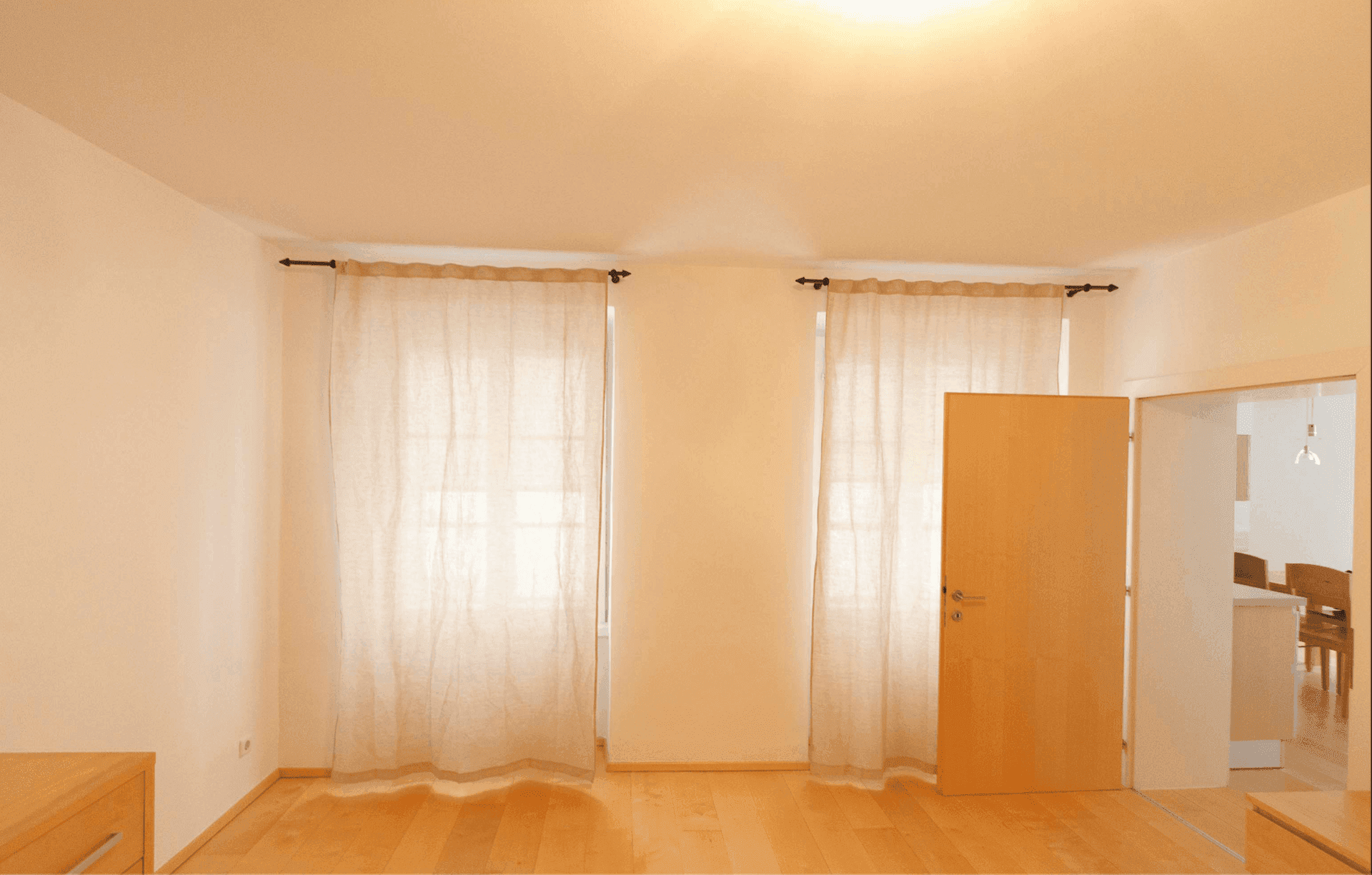 Heller Raum mit zwei Fenstern, Vorhängen und einer Holztür, die zu einem angrenzenden Essbereich führt.