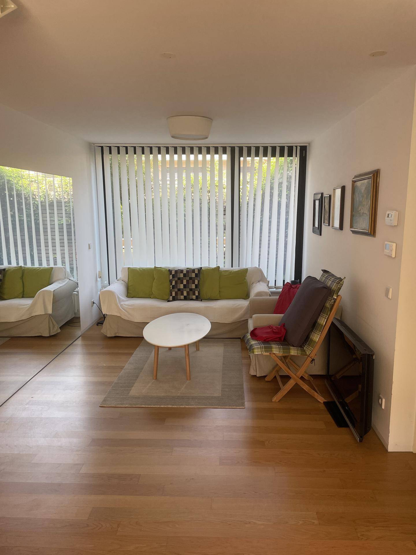 Helles Wohnzimmer mit weißem Sofa, grünen Kissen und großen Fenstern mit Jalousien.