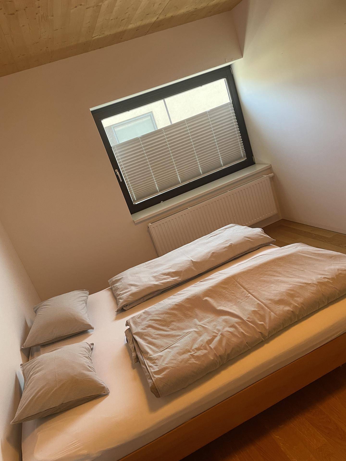 Helles Schlafzimmer mit Holzbett, schlichten Möbeln und einem Fenster mit Plissee.