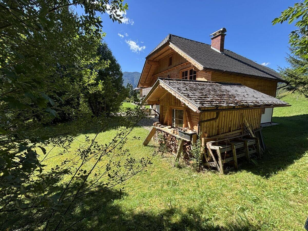 Rustikales Holzhaus mit Schuppen und Holzstapel, eingebettet in eine grüne Landschaft mit Bergblick.