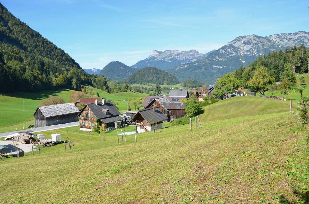 Weitläufige Ansicht eines Bergdorfes mit mehreren Holzhäusern, grünen Hängen und einer beeindruckenden Bergkette.