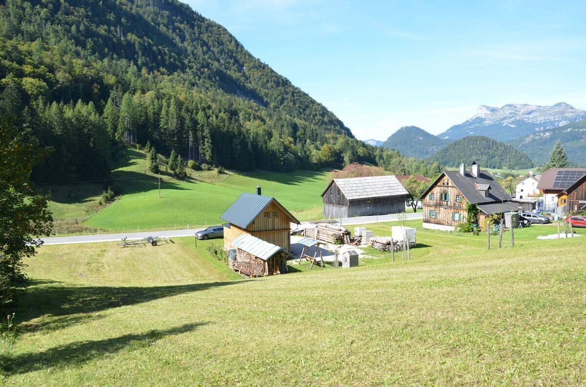 Weitläufige ländliche Ansicht mit mehreren Holzhäusern, grünen Wiesen und majestätischen Bergen im Hintergrund.