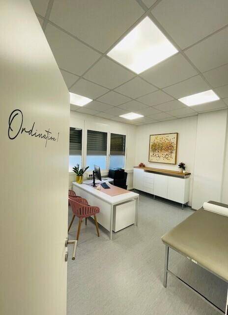 Helles Büro mit Schreibtisch, Untersuchungsbank und Fenster mit Jalousien für Privatsphäre.