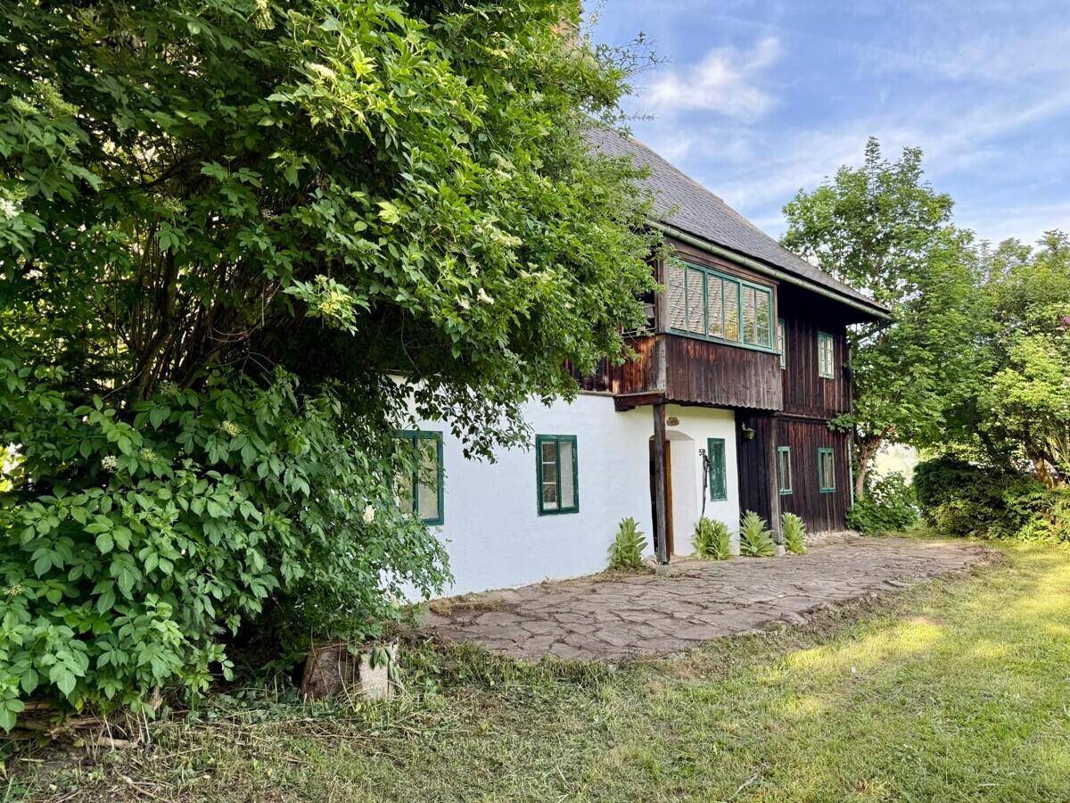 Seitlicher Blick auf das Landhaus mit rustikaler Steinterrasse und üppiger Vegetation im Garten.