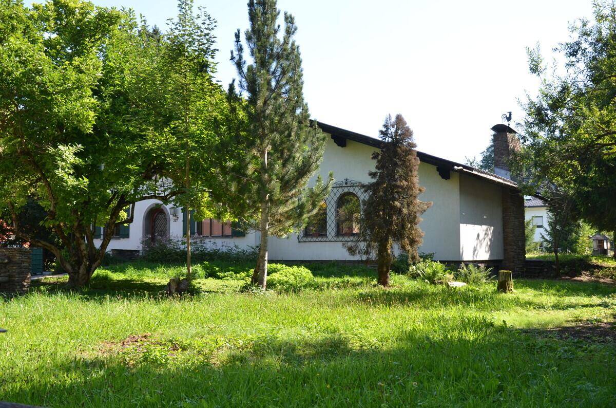 Haus umgeben von viel Grün, mit Bäumen und einer weiten Rasenfläche.
