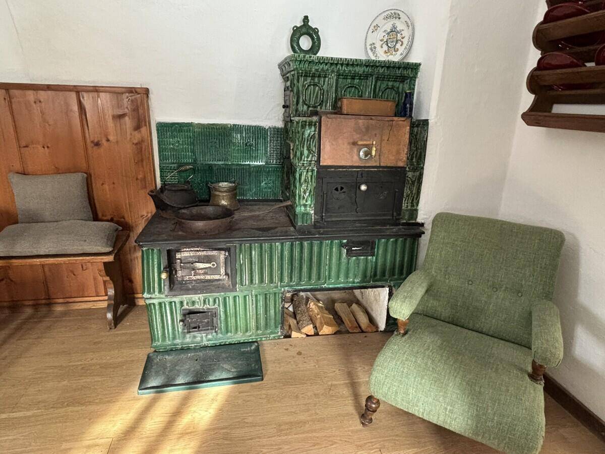 Detailansicht eines traditionellen grünen Kachelofens mit Holzscheiten und einem Sessel.