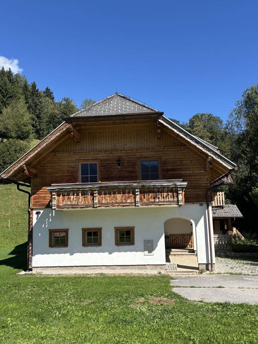 Charmantes Holzhaus mit Satteldach und Holzbalkonen, umgeben von grünen Hügeln und Bäumen.