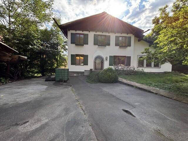 Traditionelles Haus mit weißer Fassade, grünen Fensterläden und großem Vorplatz umgeben von Bäumen.