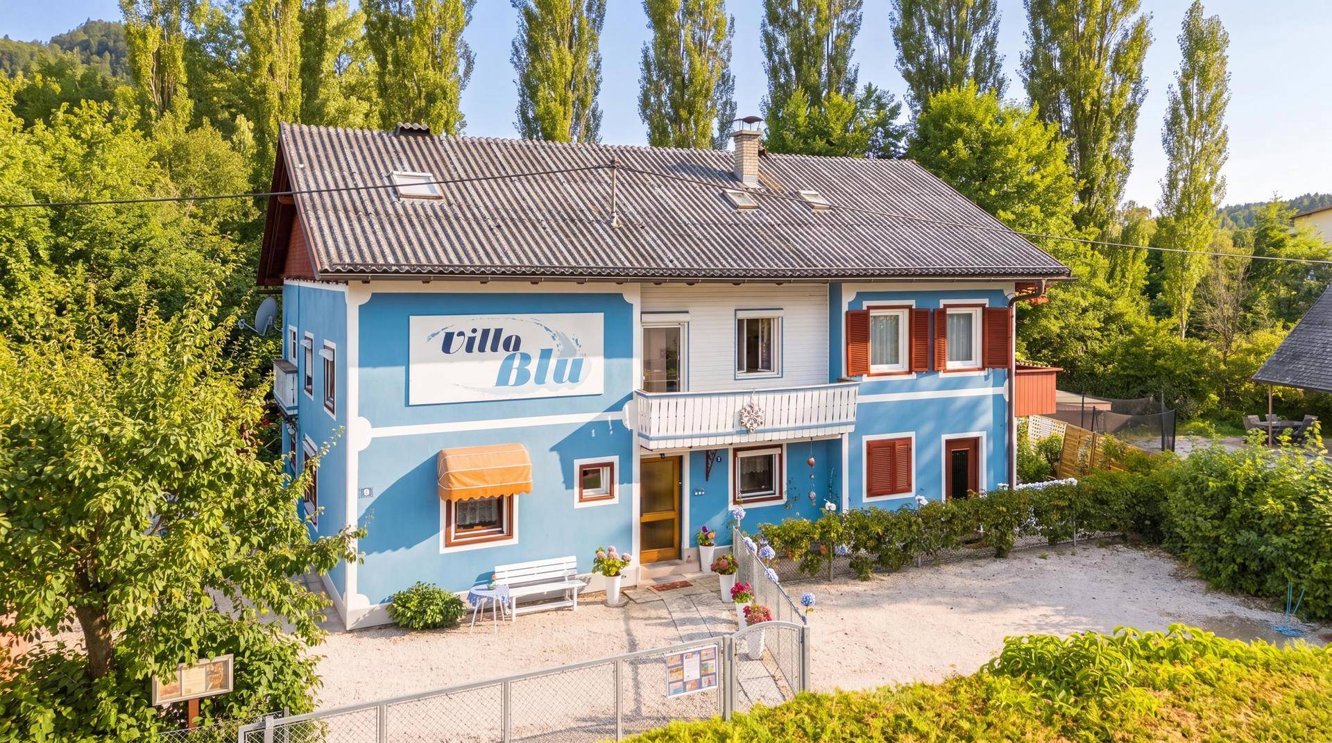 Charmantes blau-weißes Haus mit Satteldach, umgeben von hohen Bäumen und einem kleinen Vorgarten.